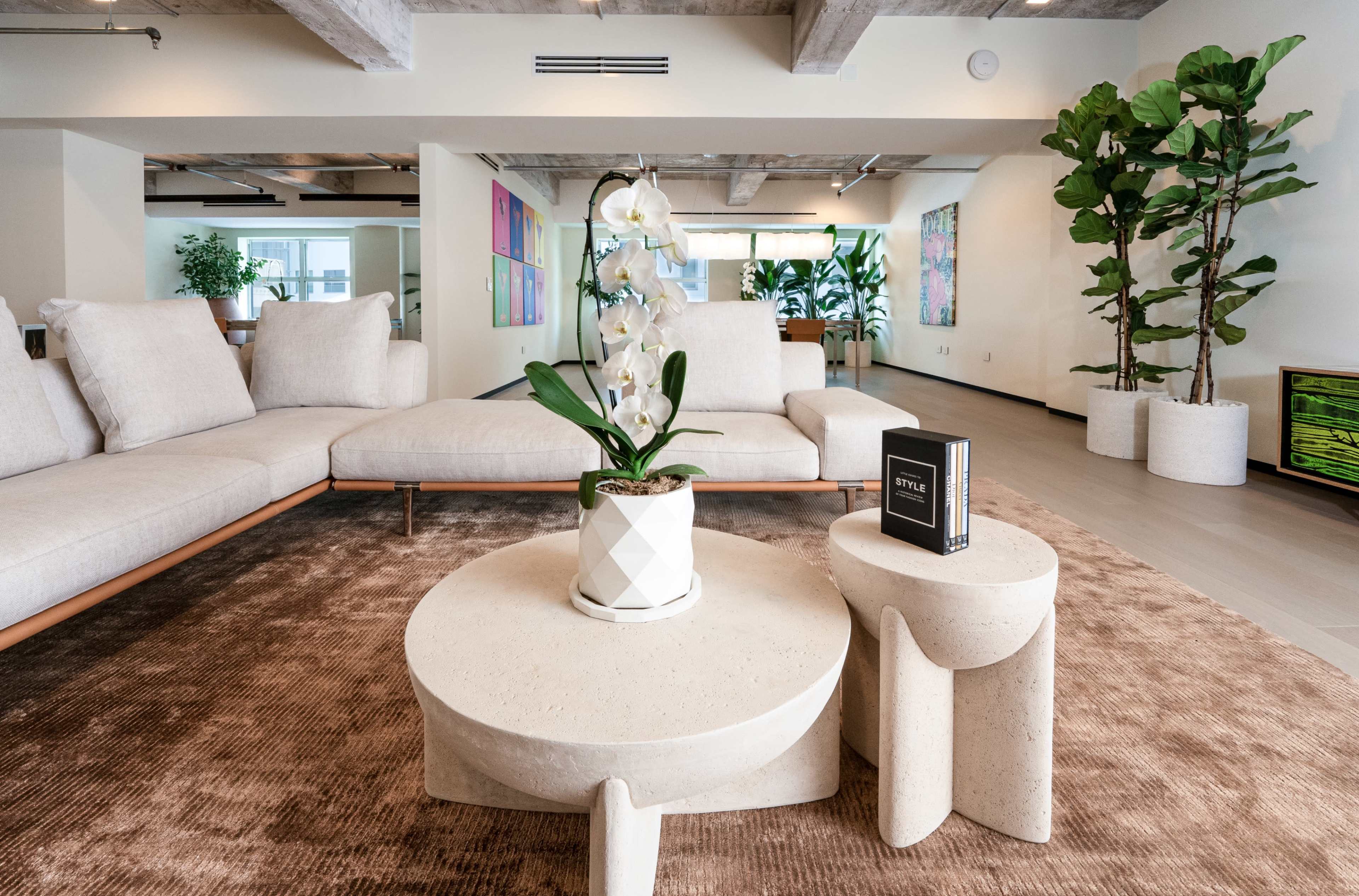 The image shows a modern living area featuring a large sectional sofa, a round coffee table with a plant, and decorative elements including art on the walls and potted plants.
