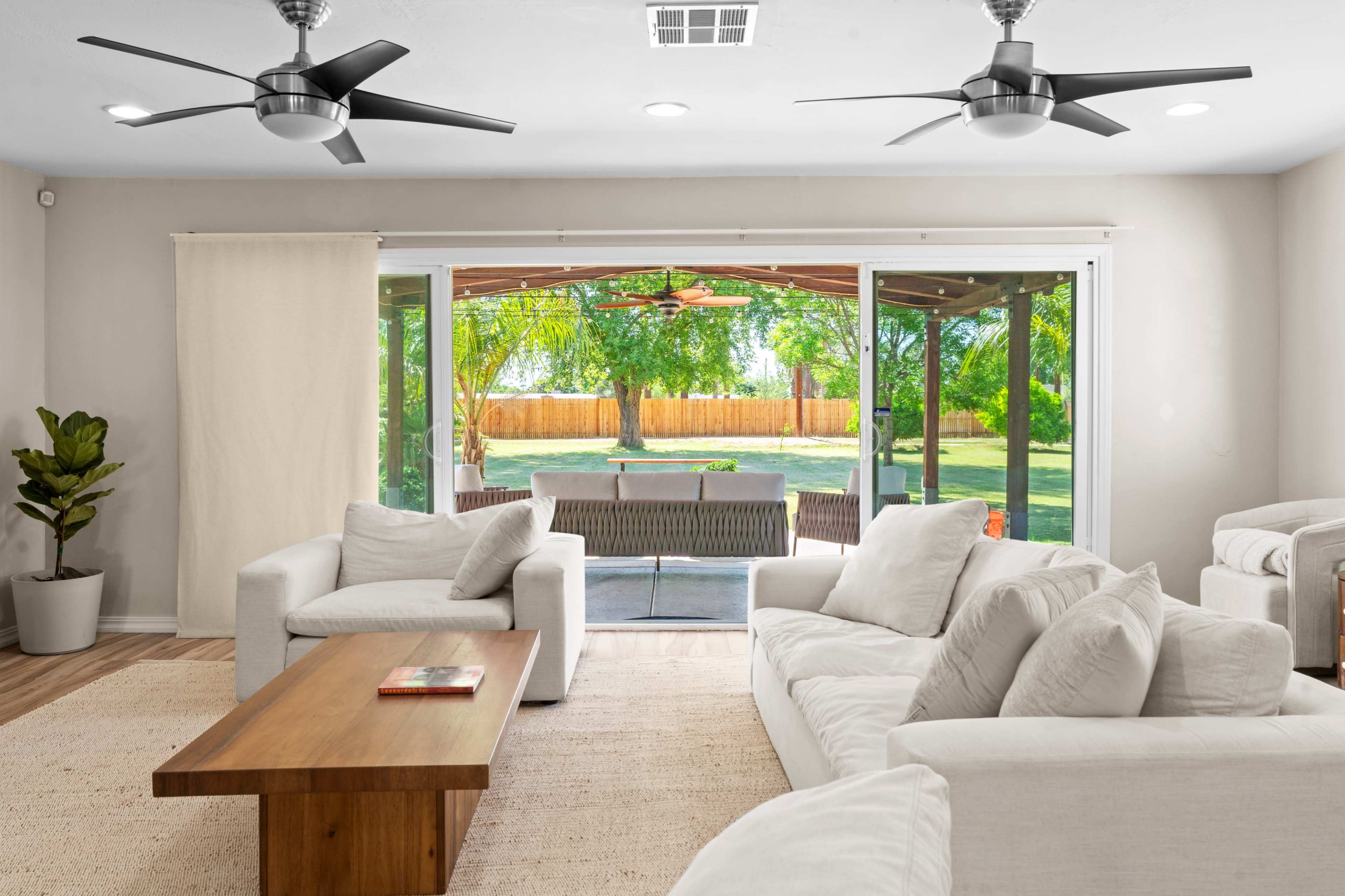 A modern living room features white furniture, a wooden coffee table, and large glass doors that open to a backyard with a shaded seating area.