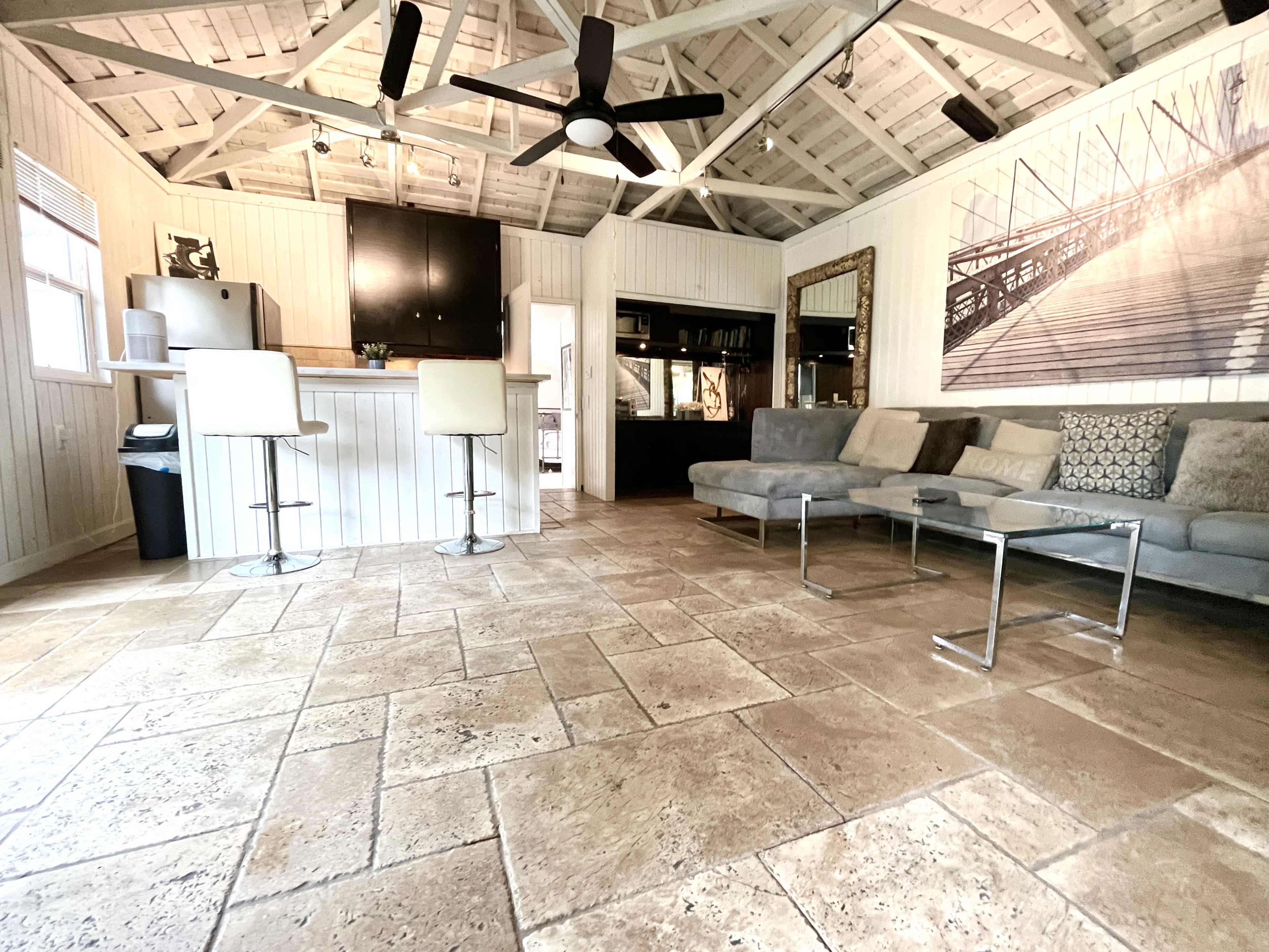 The image shows a modern living space with a bar area, a sectional sofa, and stone flooring beneath a vaulted ceiling with exposed beams.