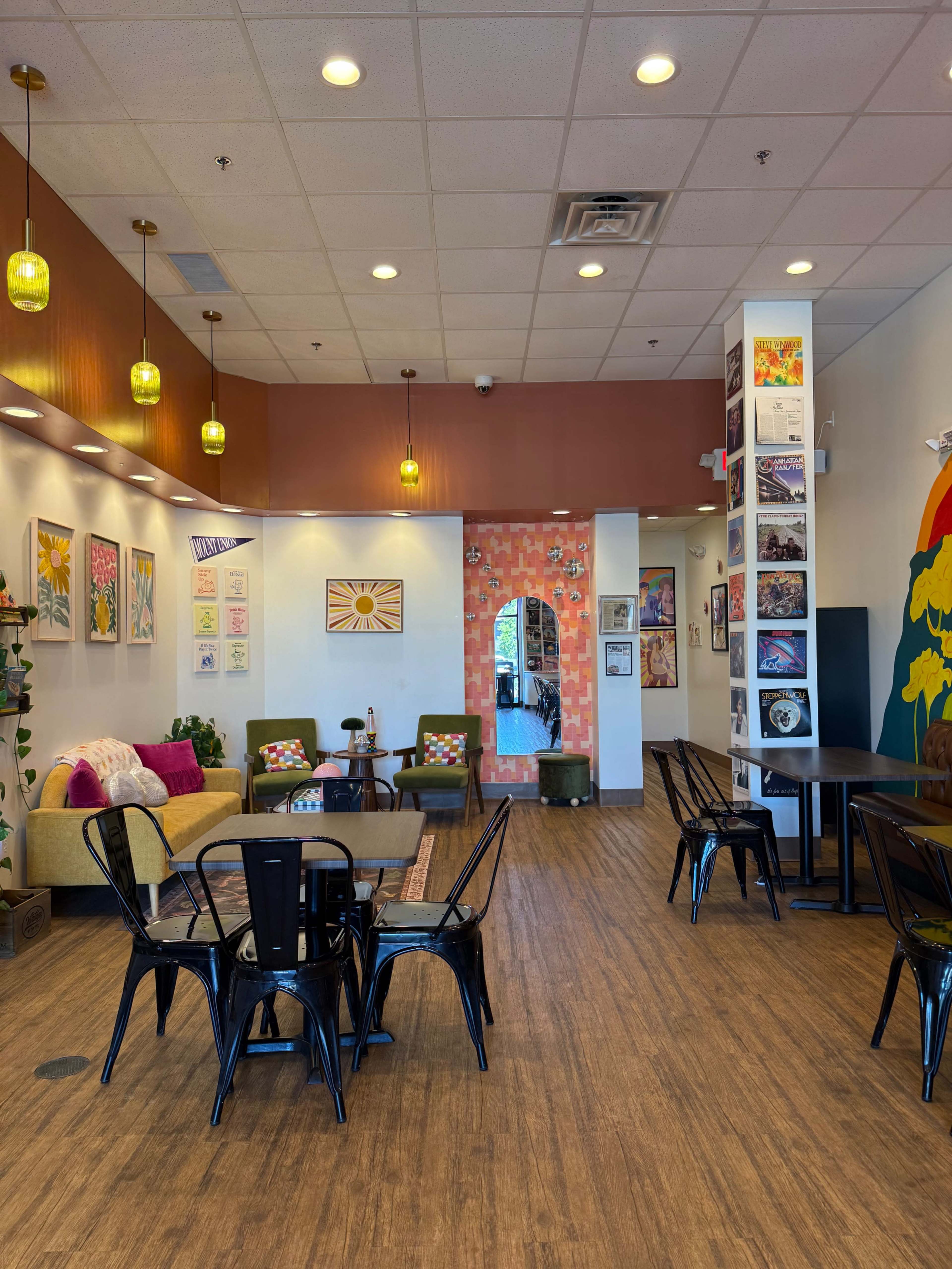The image shows a brightly lit café interior with colorful seating, art on the walls, and wooden flooring.