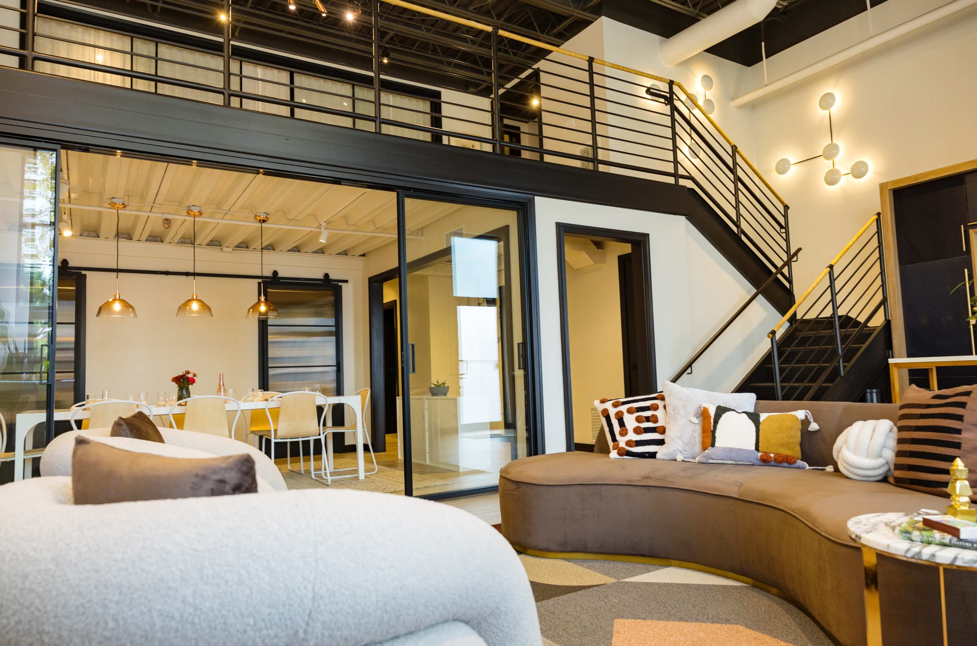 A modern, open-concept interior with a curved sofa, a dining area with white chairs, and a staircase leading to an upper level.