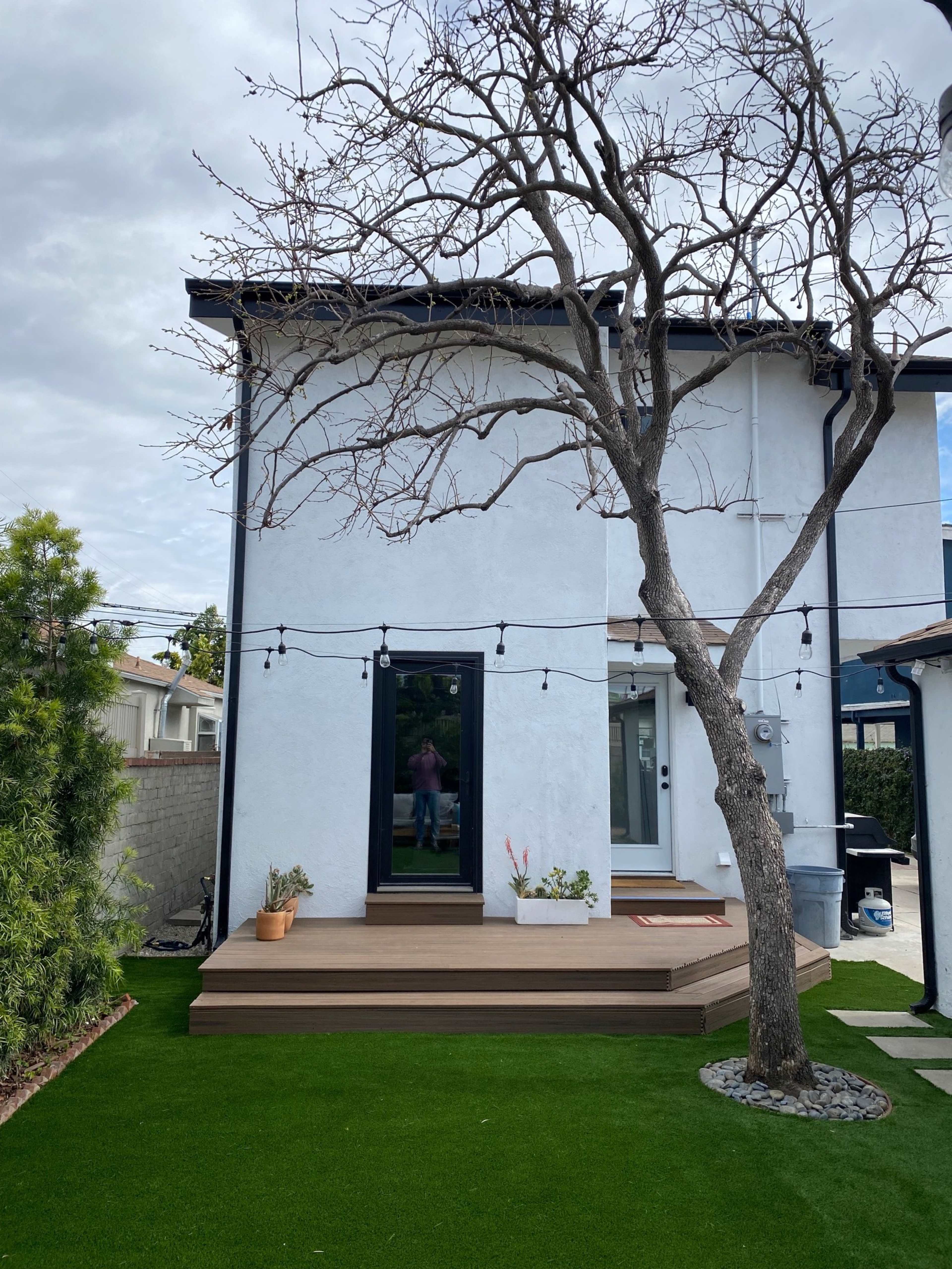 The image shows a modern two-story white house with a deck, a bare tree, and green artificial grass in the foreground.