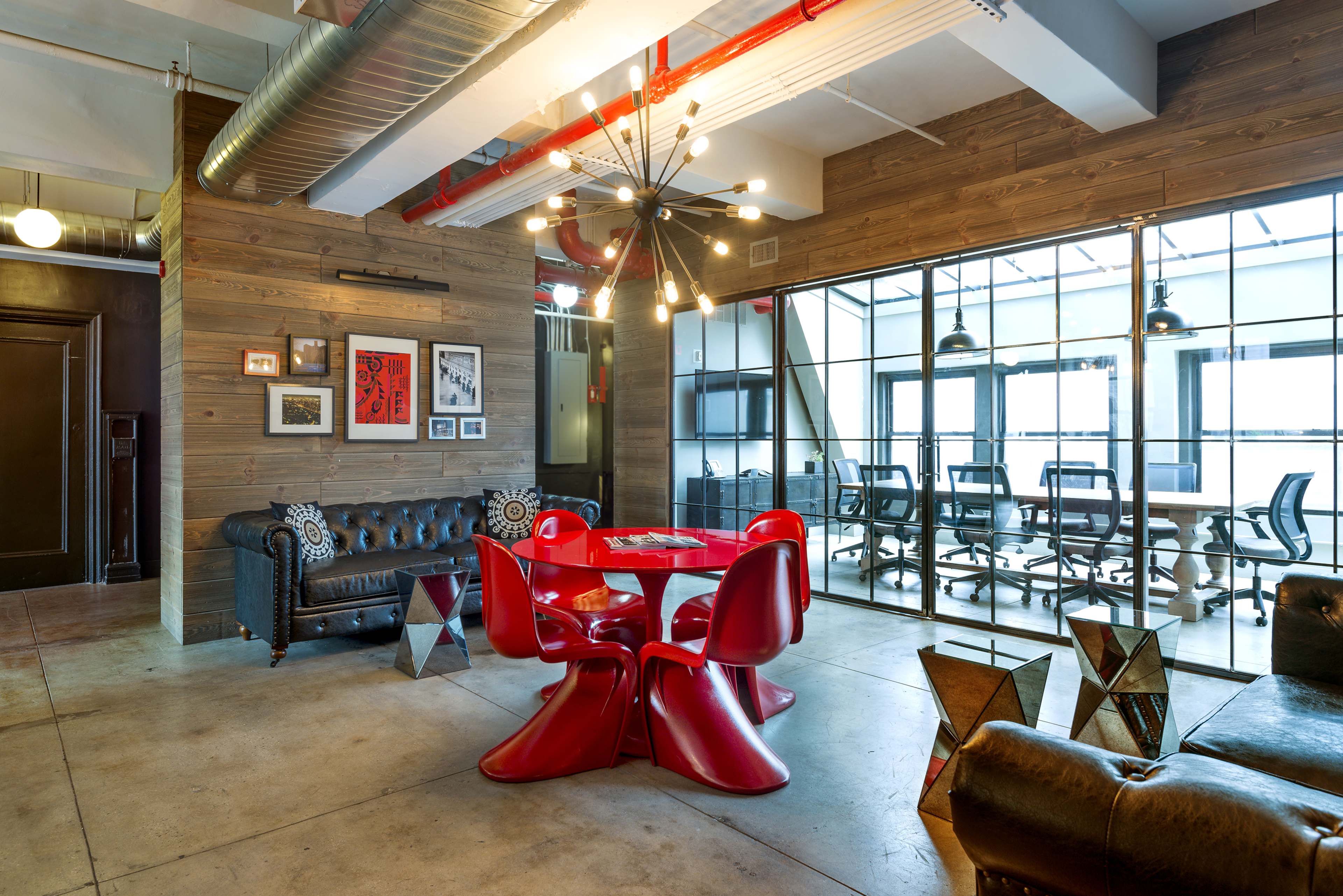 A modern office space features a red table surrounded by red chairs, with a seating area of black leather sofas and a backdrop of wood-paneled walls.