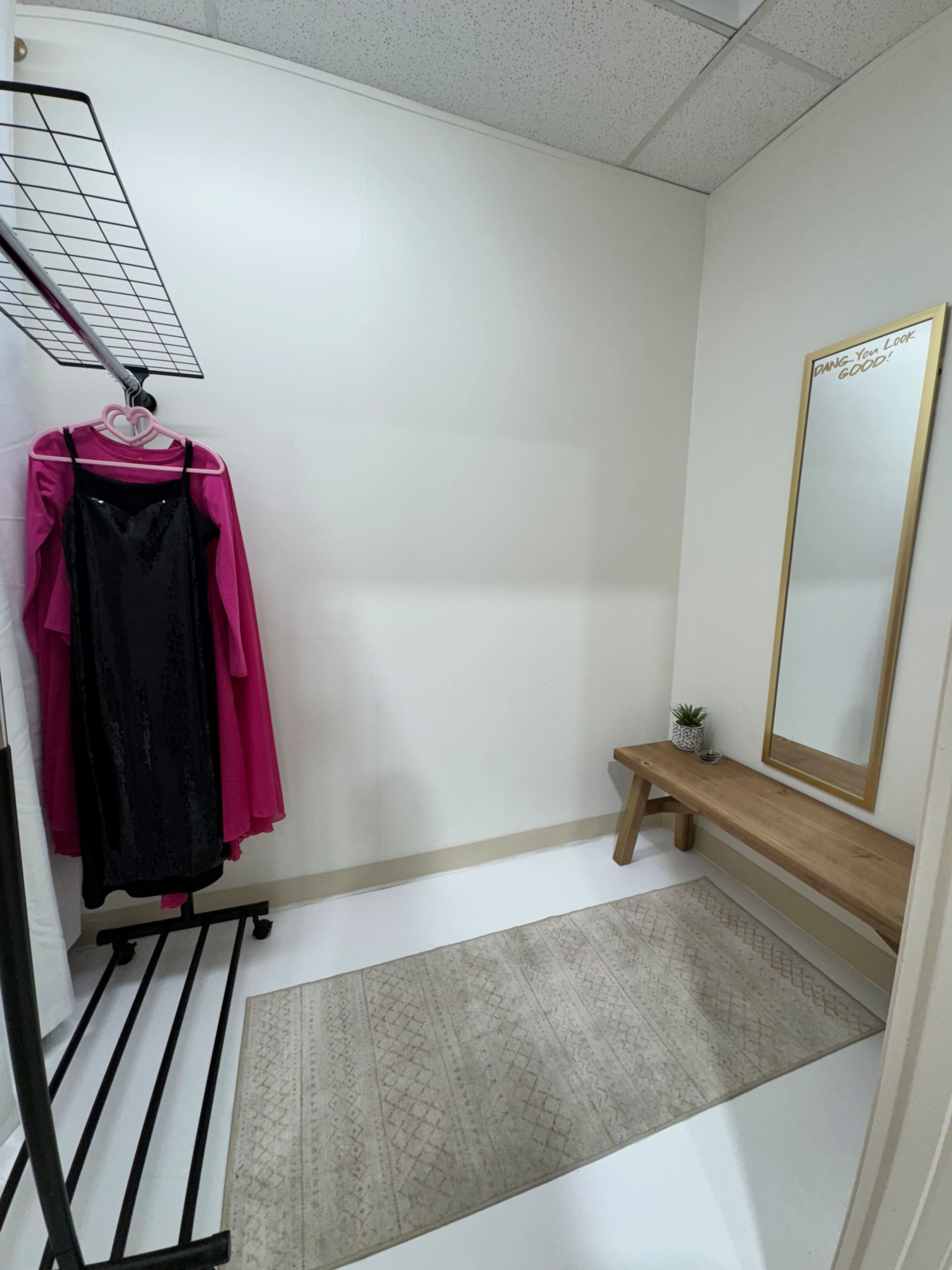 The image shows a fitting room with a mirror, a wooden bench, a rug, and clothing hung on a rack.