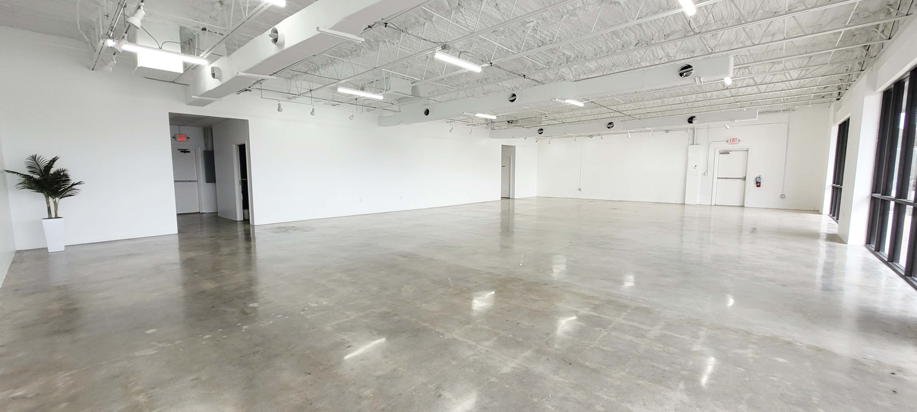 A spacious, empty room with polished concrete floors and white walls, featuring several light fixtures and doors along the perimeter.