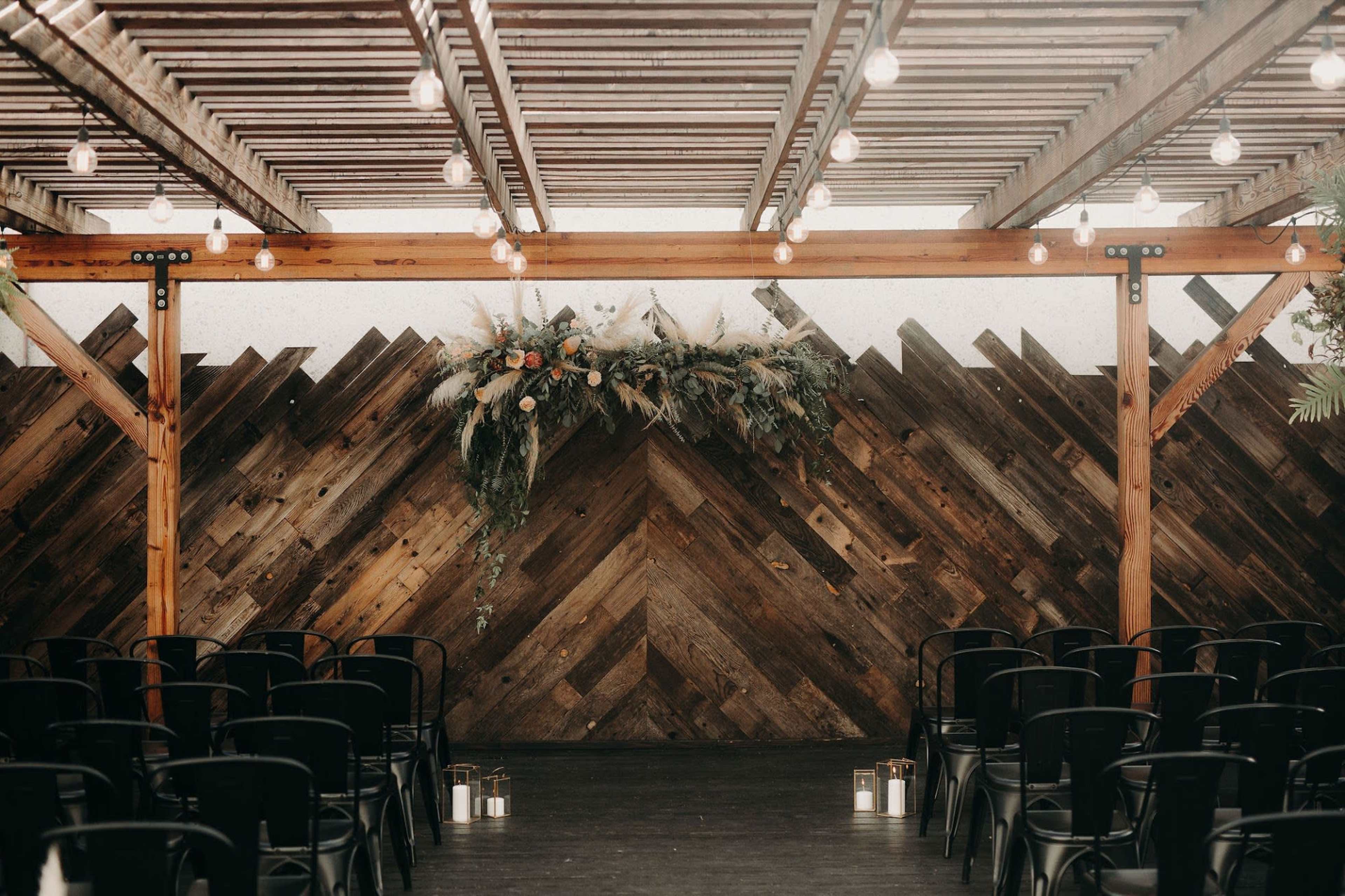 The image shows a decorated indoor venue with wooden beams, a rustic wooden backdrop, and rows of black chairs facing a floral arrangement hanging above.