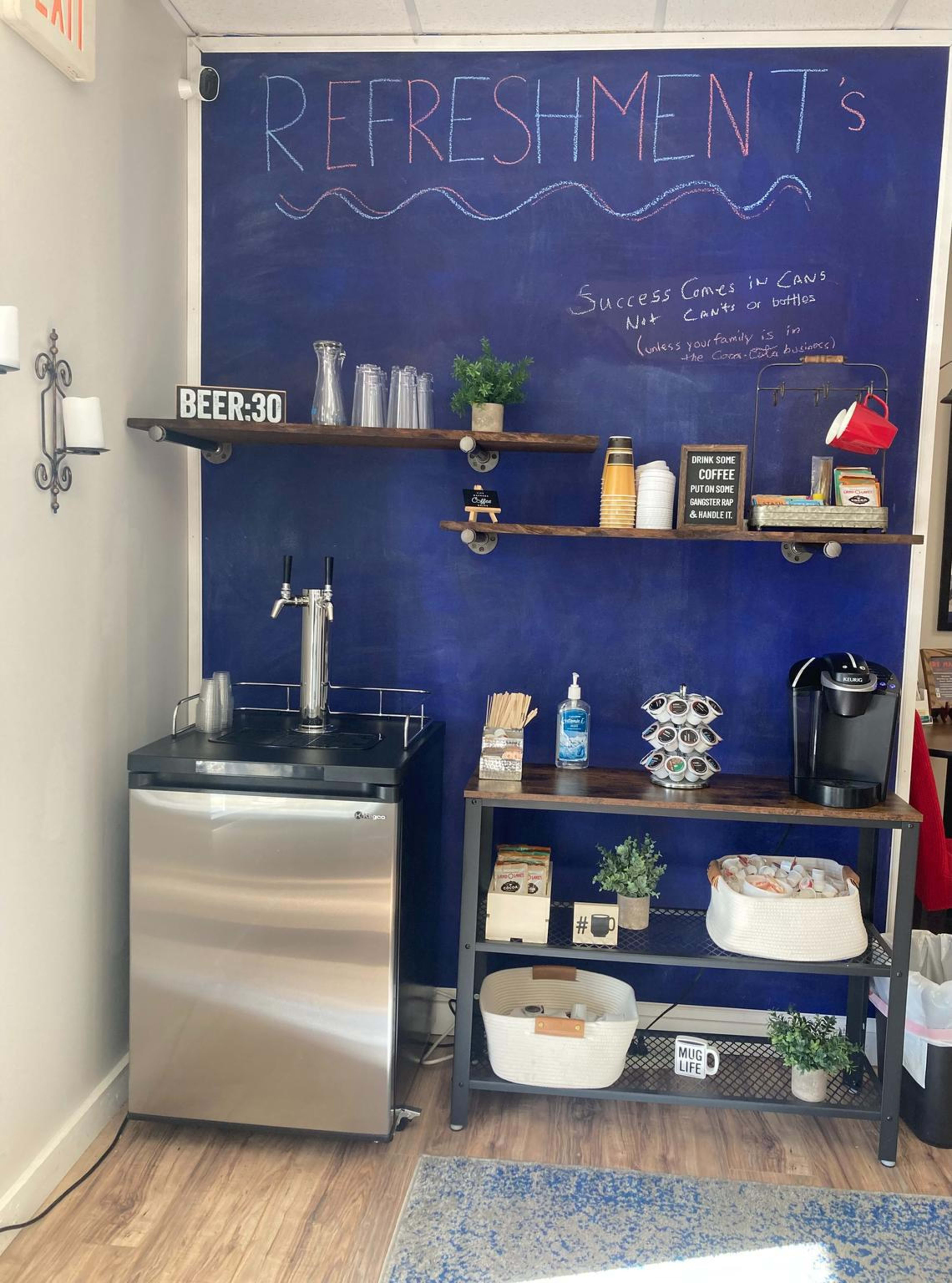 A refreshment station with a small refrigerator, shelves holding various cups and snacks, a coffee maker, and a chalkboard wall outlining the word "REFRESHMENTS" and a motivational quote.