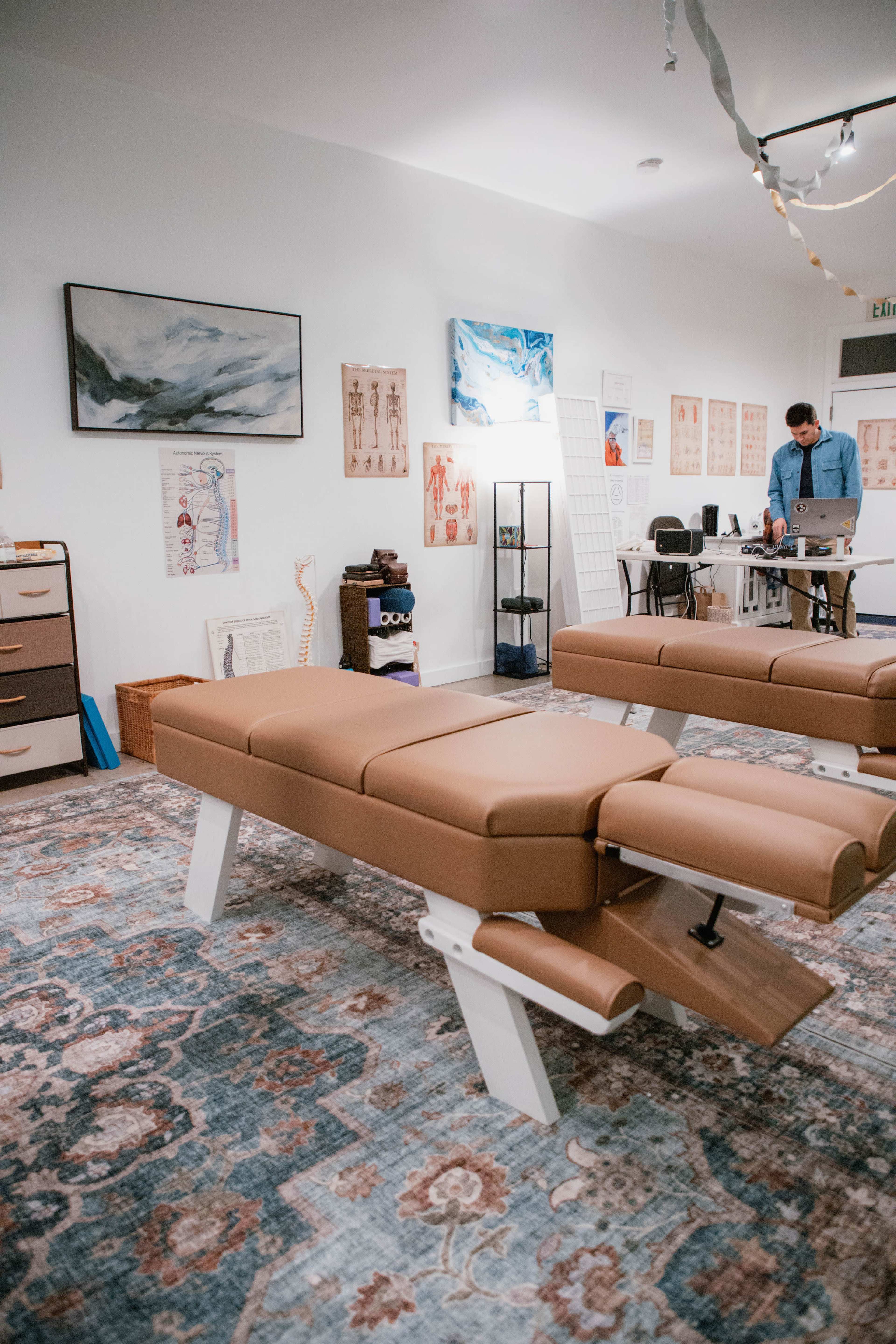 The image shows a massage therapy room with a brown treatment table, anatomical posters on the walls, and a person working on a laptop in the background.