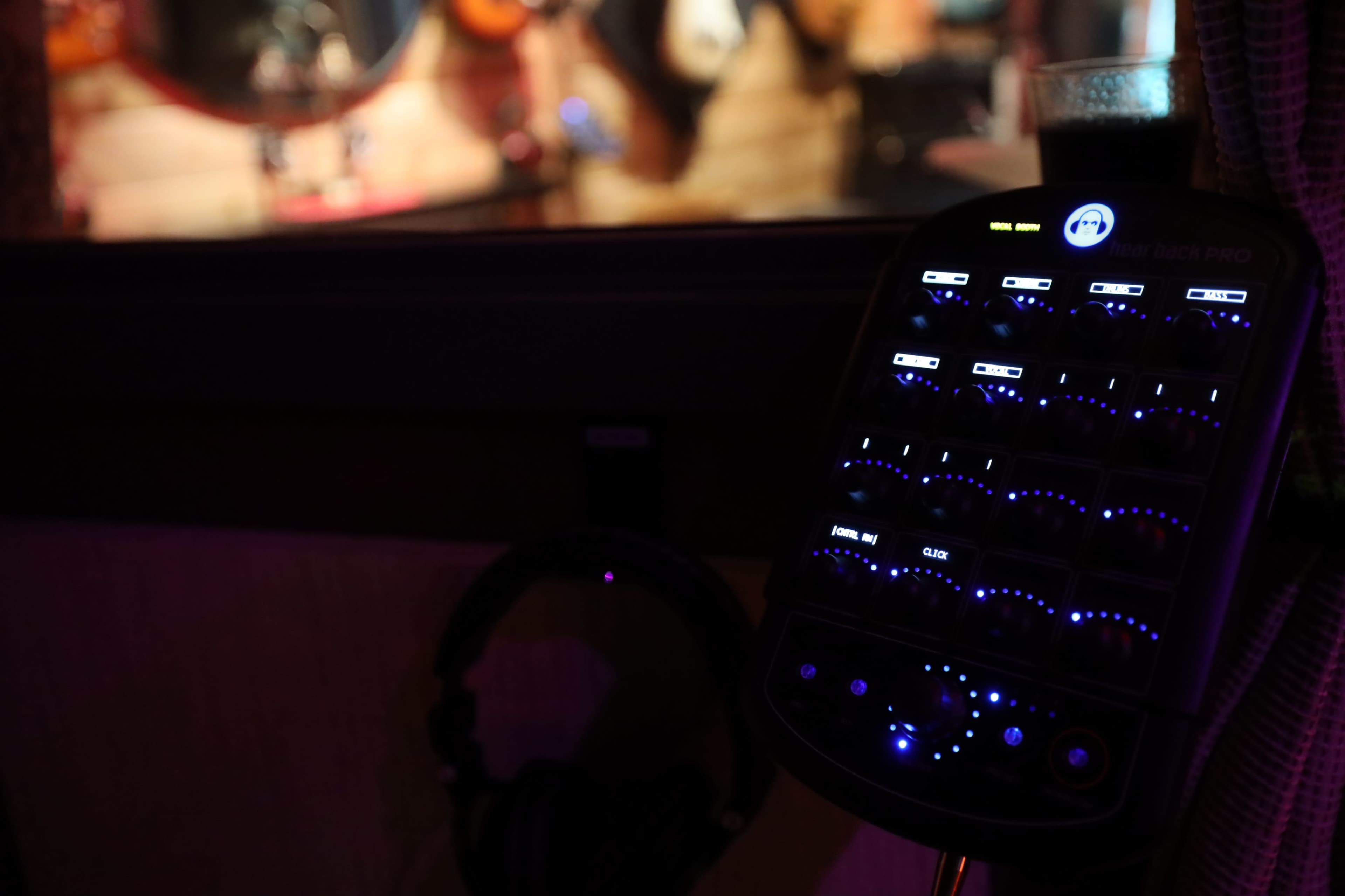 A digital mixer with illuminated controls is positioned in the foreground, with a blurred backdrop of musicians performing in a studio.