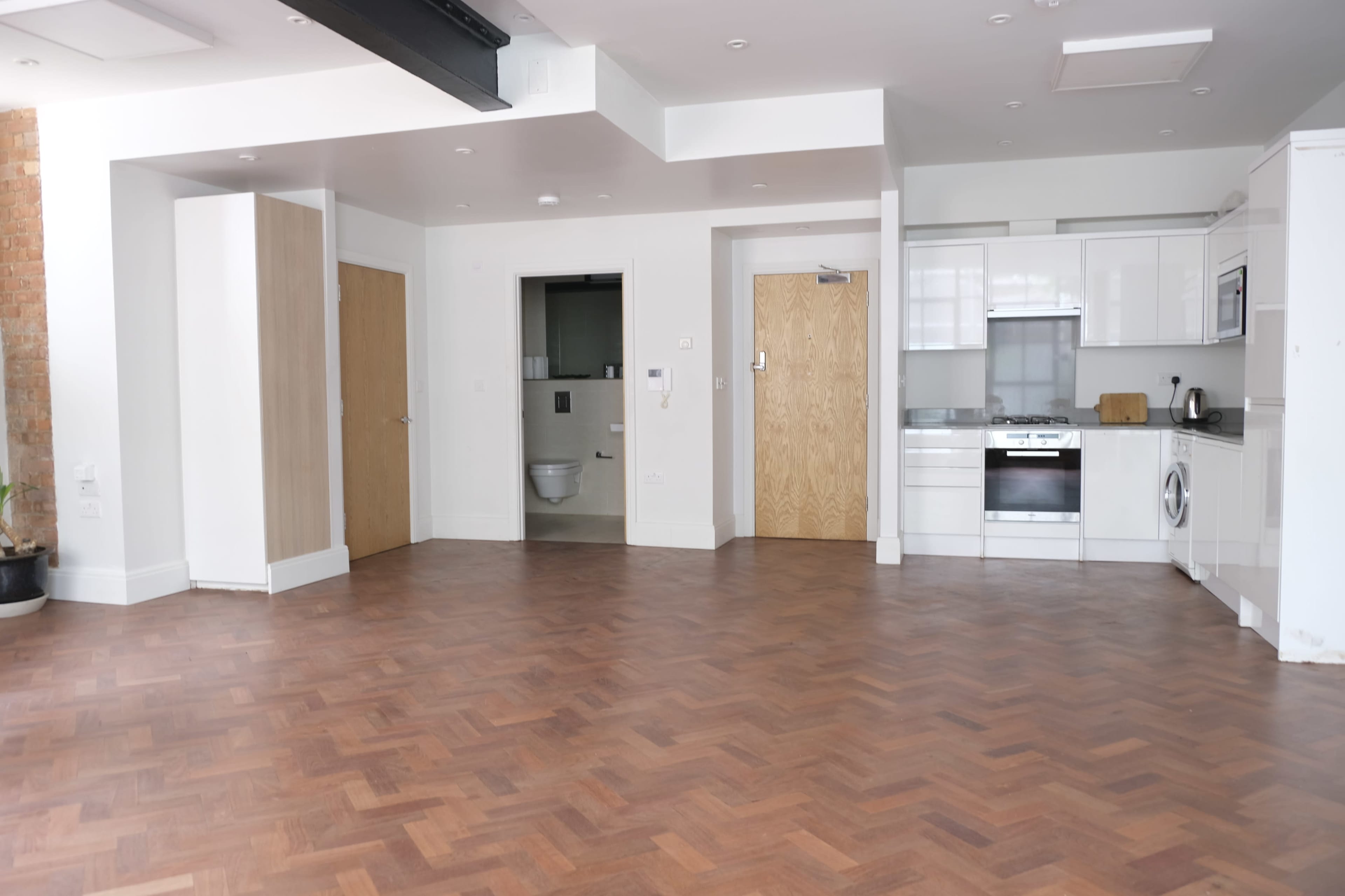 An empty room with wooden flooring, featuring a kitchenette and doorways leading to a bathroom and another room.