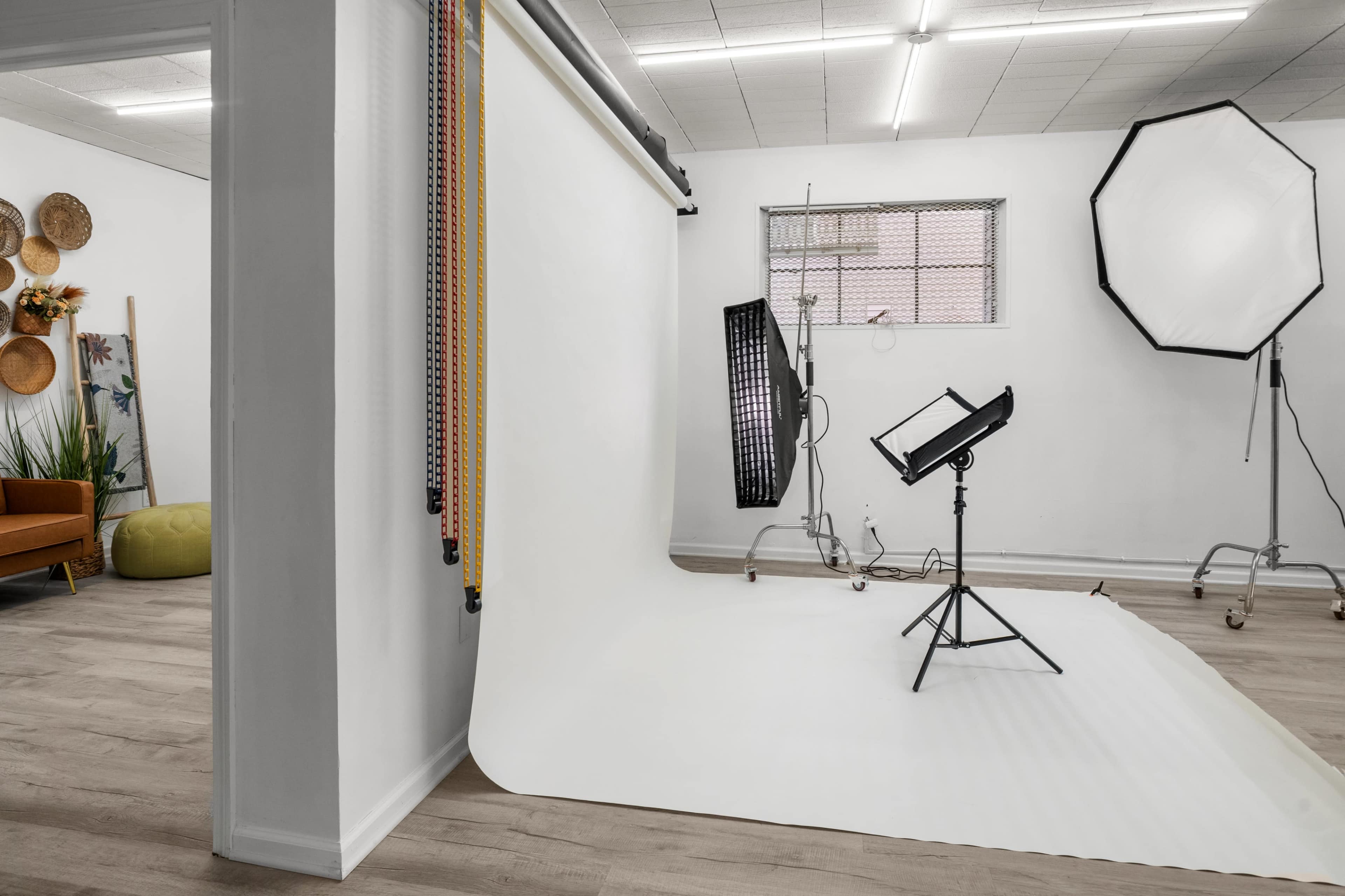 The image shows a photography studio with a white backdrop, lighting equipment, and soft furnishings.