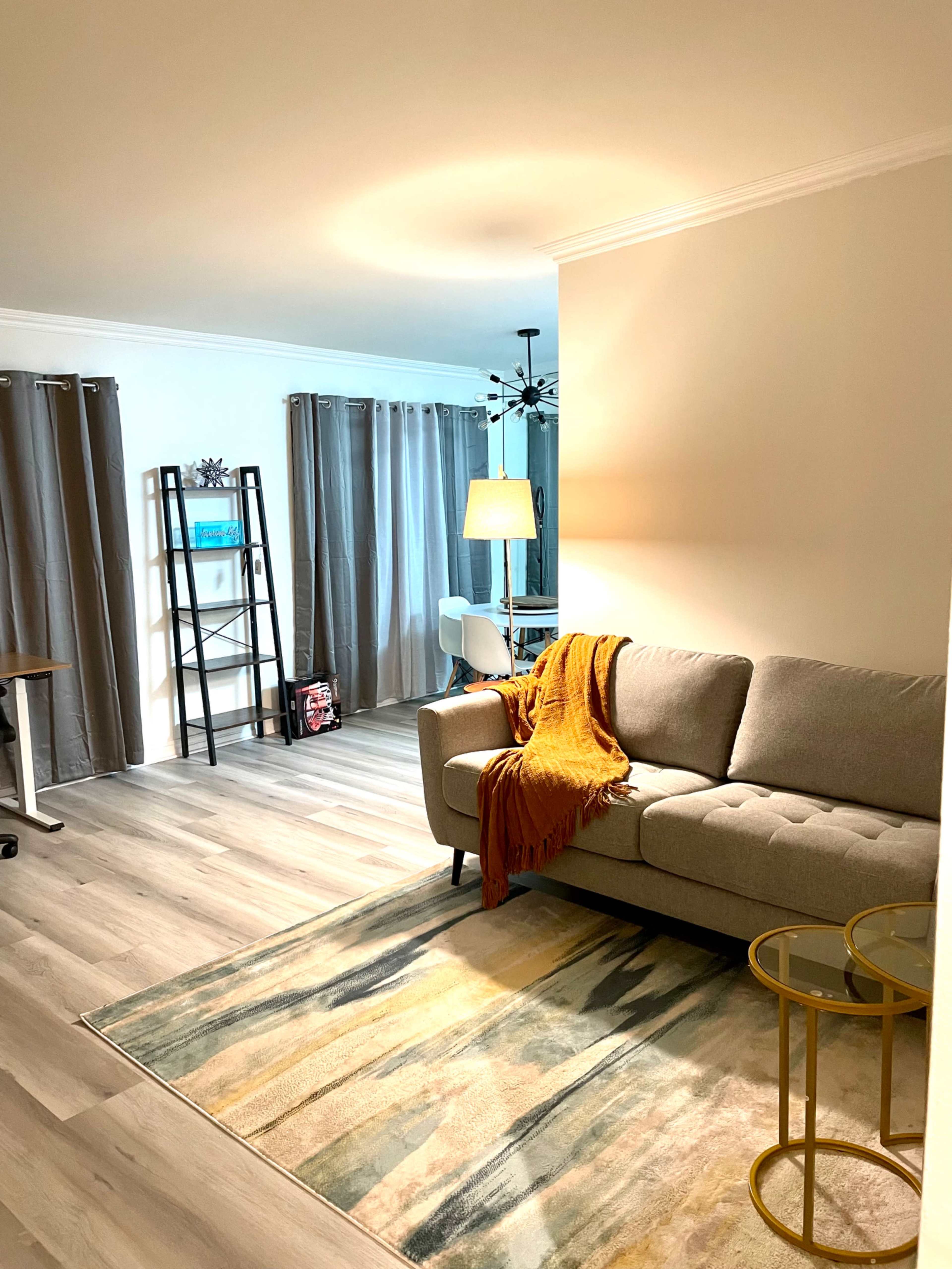A neatly arranged living room with a gray sofa, a mustard-colored throw blanket draped over it, a patterned area rug, and a tall bookshelf against the wall.