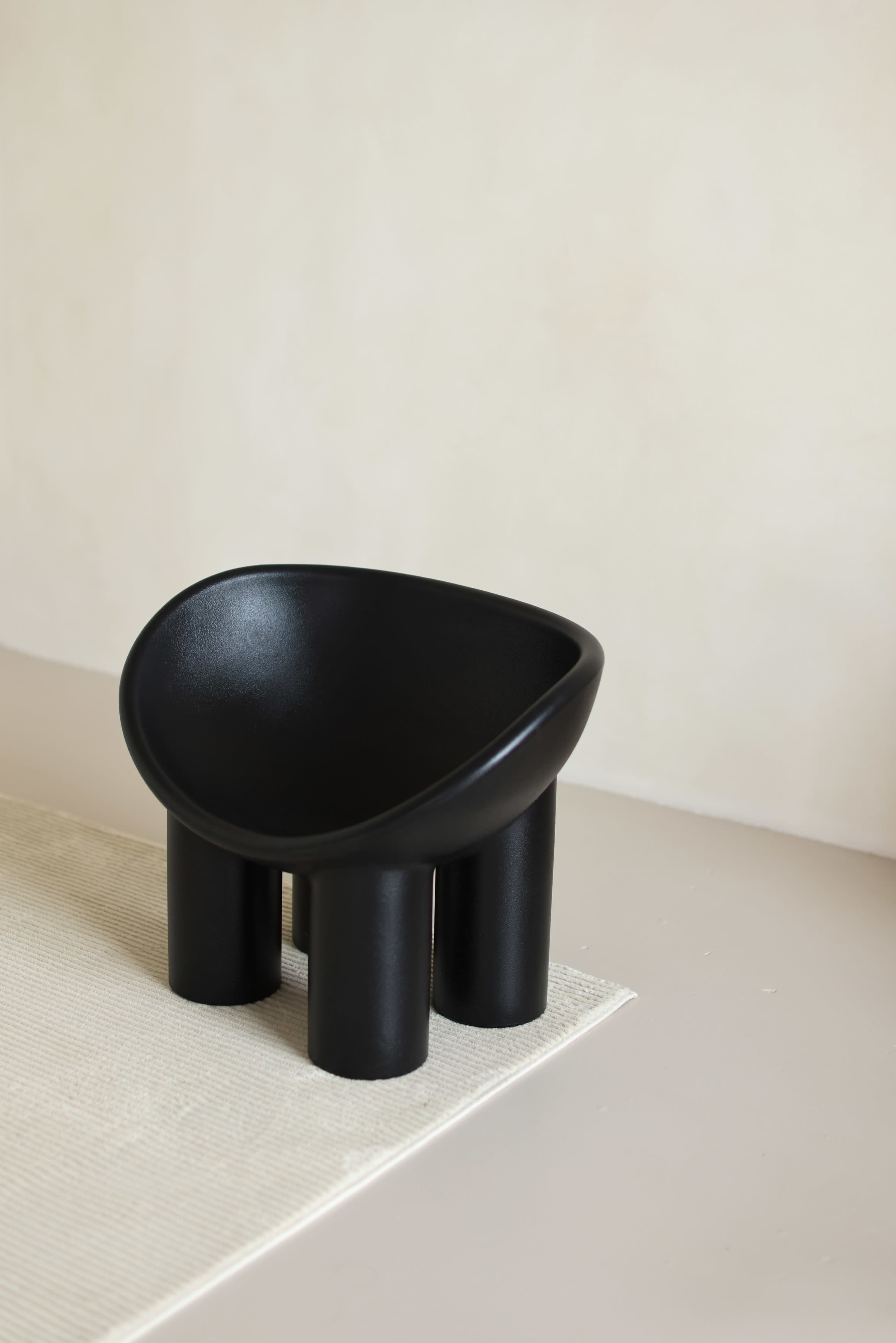 A black modern chair with a curved seat and four cylindrical legs sits on a light-colored surface next to a neutral wall.