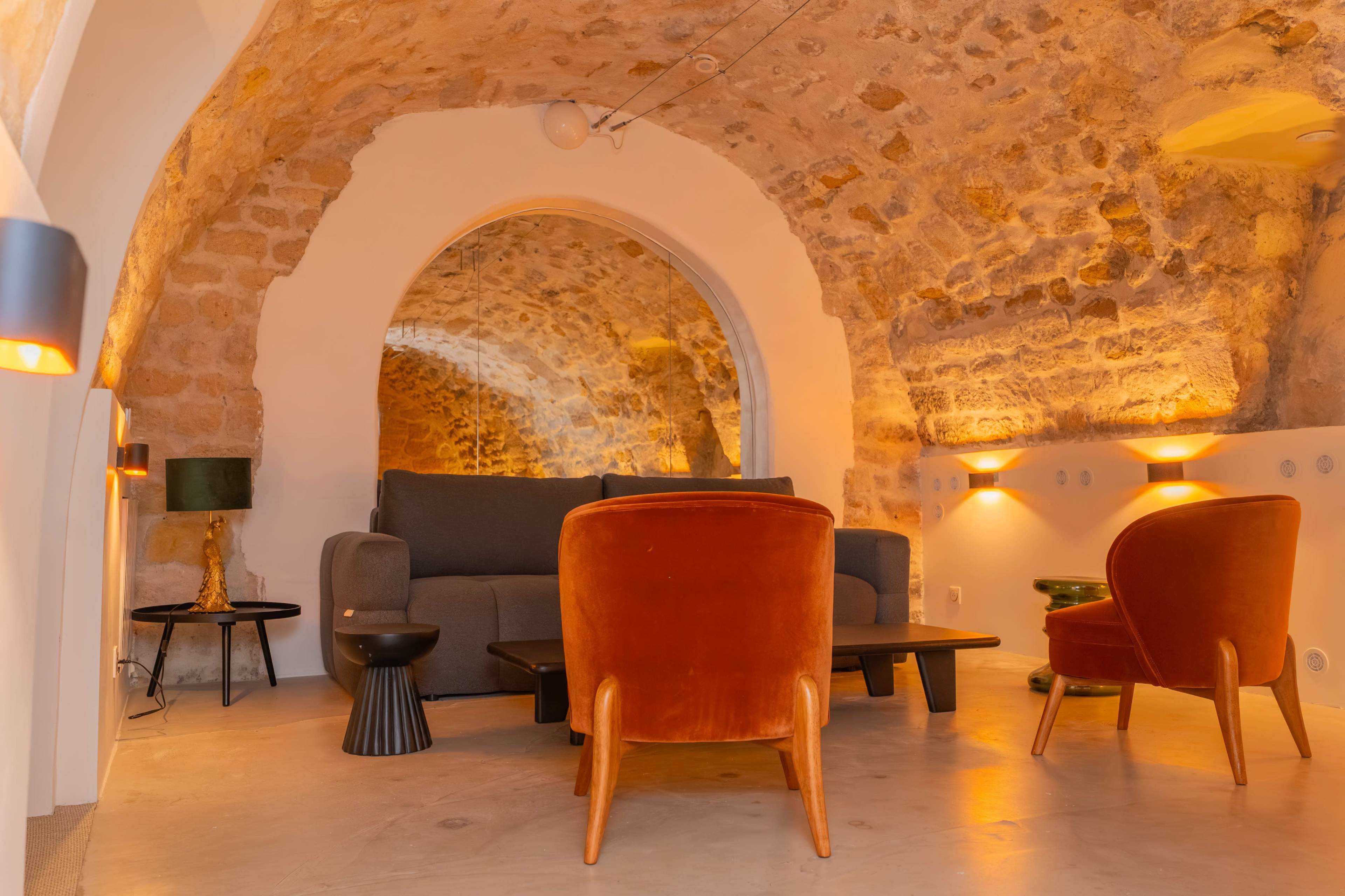 The image shows a cozy lounge area with stone walls, featuring a grey sofa, two red armchairs, a coffee table, and stylish lighting.