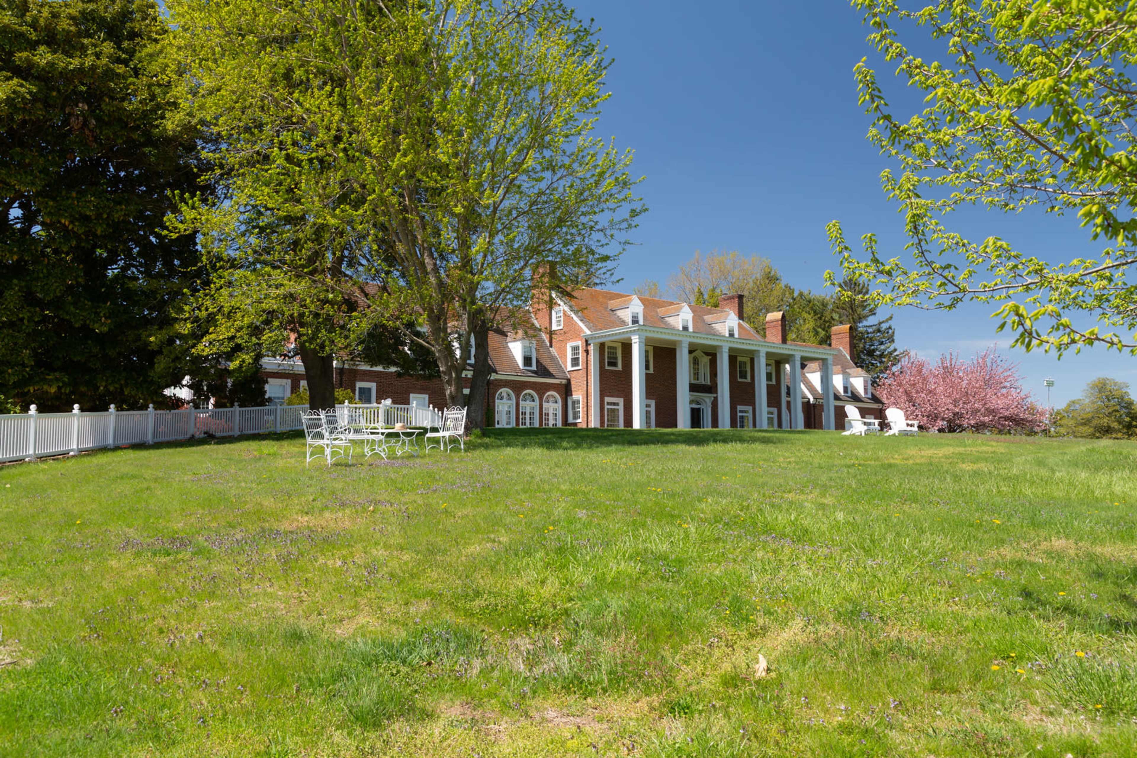 A large brick house with white columns sits on a grassy hillside, surrounded by trees and flowering plants.