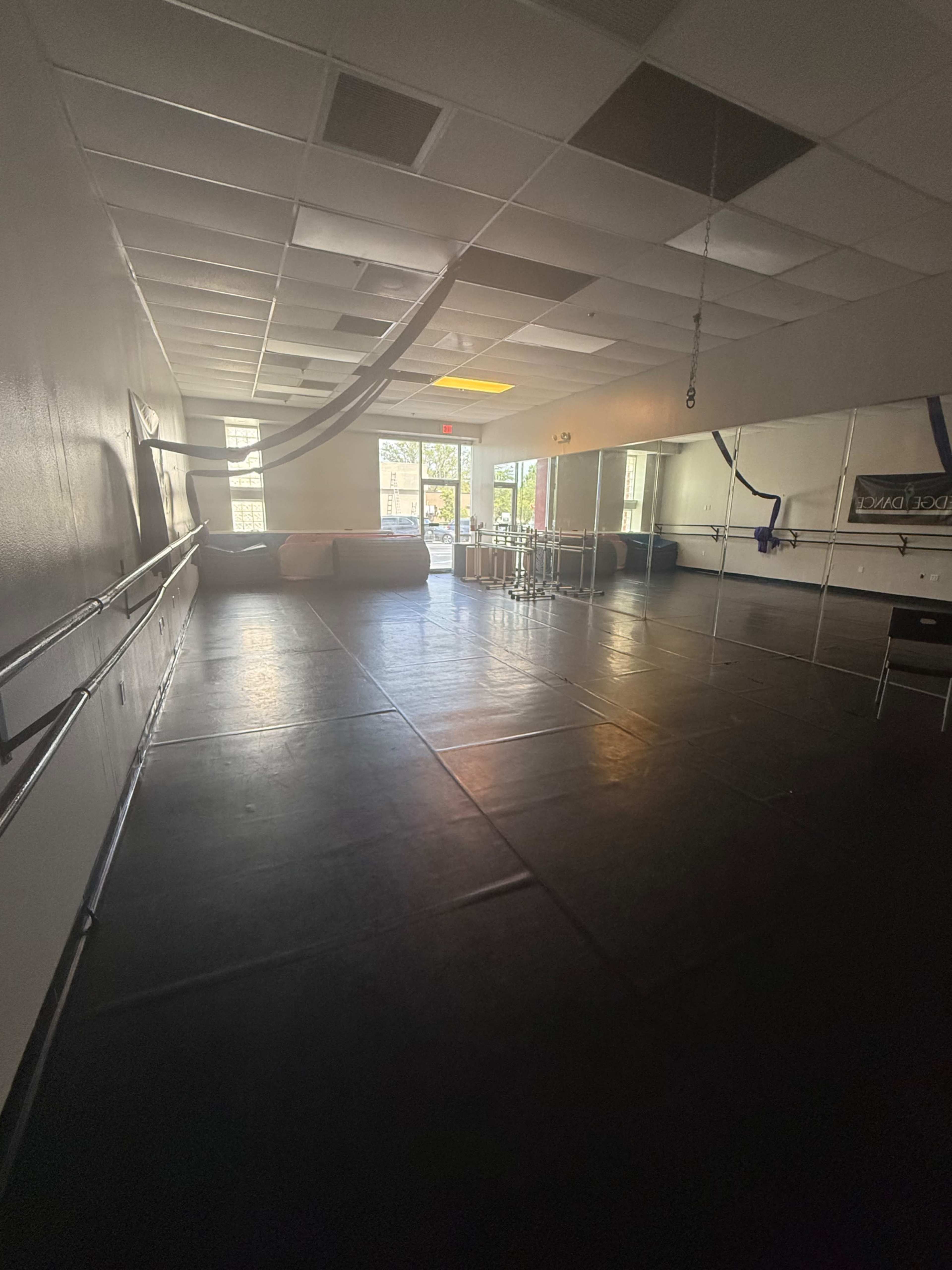 The image shows an empty dance studio with mirrored walls, a black floor, and natural light coming through windows.