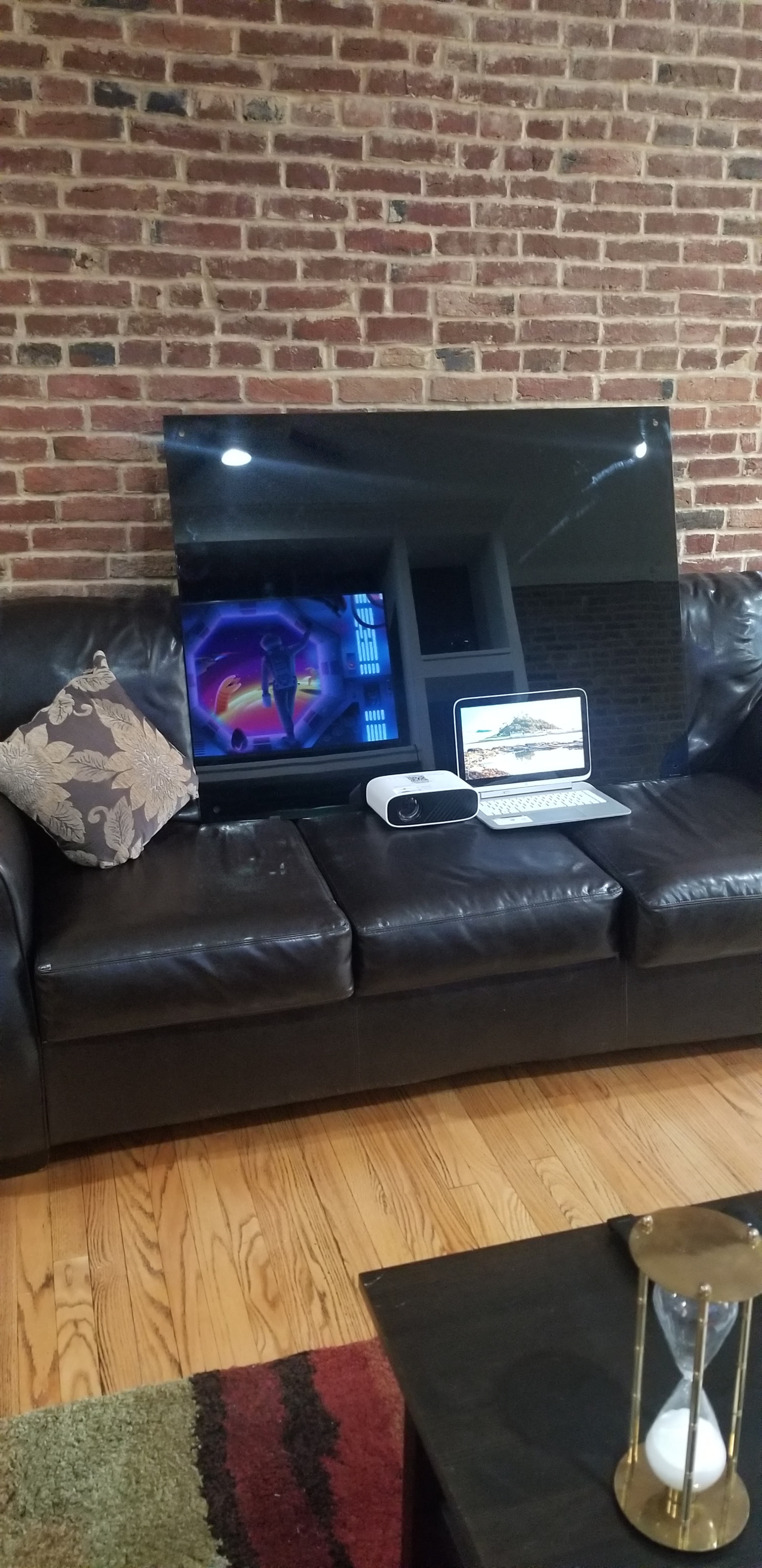 A large television is positioned on a black leather couch, accompanied by a laptop and a projector on the table in front of it, against a backdrop of exposed brick walls.
