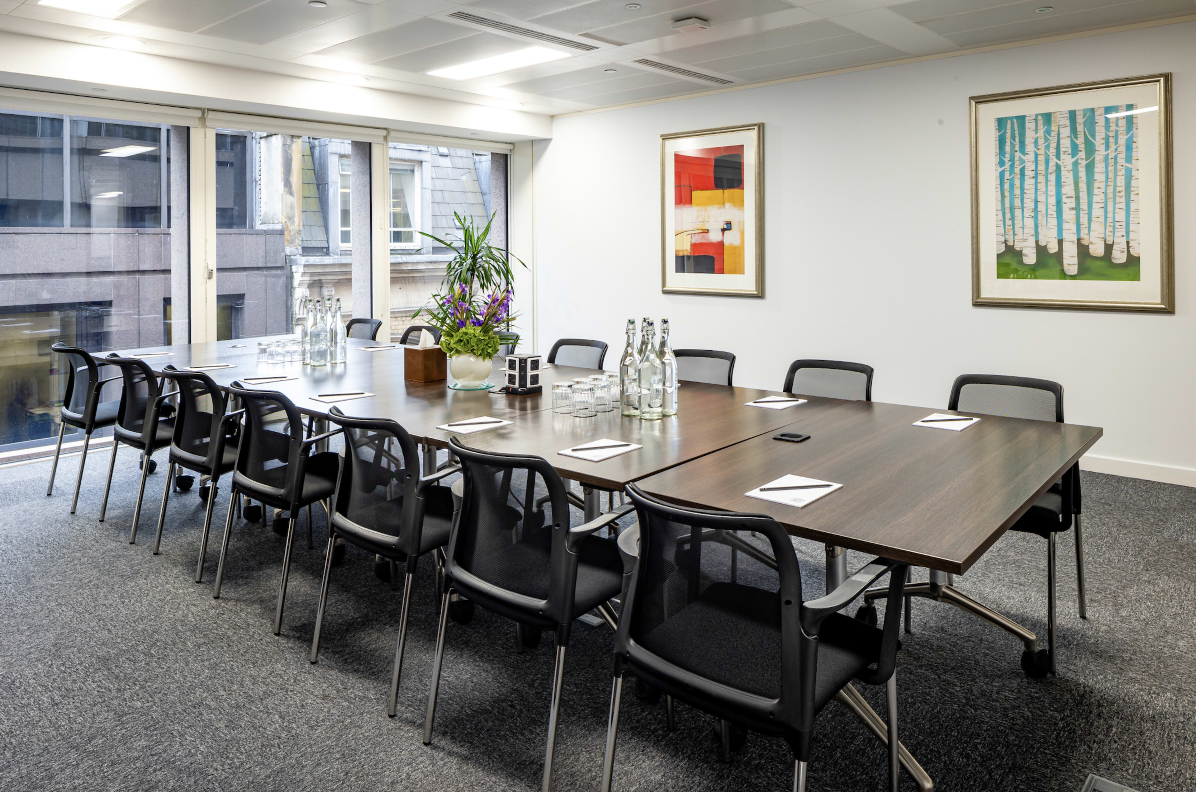 A large, modern conference room is set up for a meeting, with a long table surrounded by black chairs, glass water bottles, and artwork on the walls.