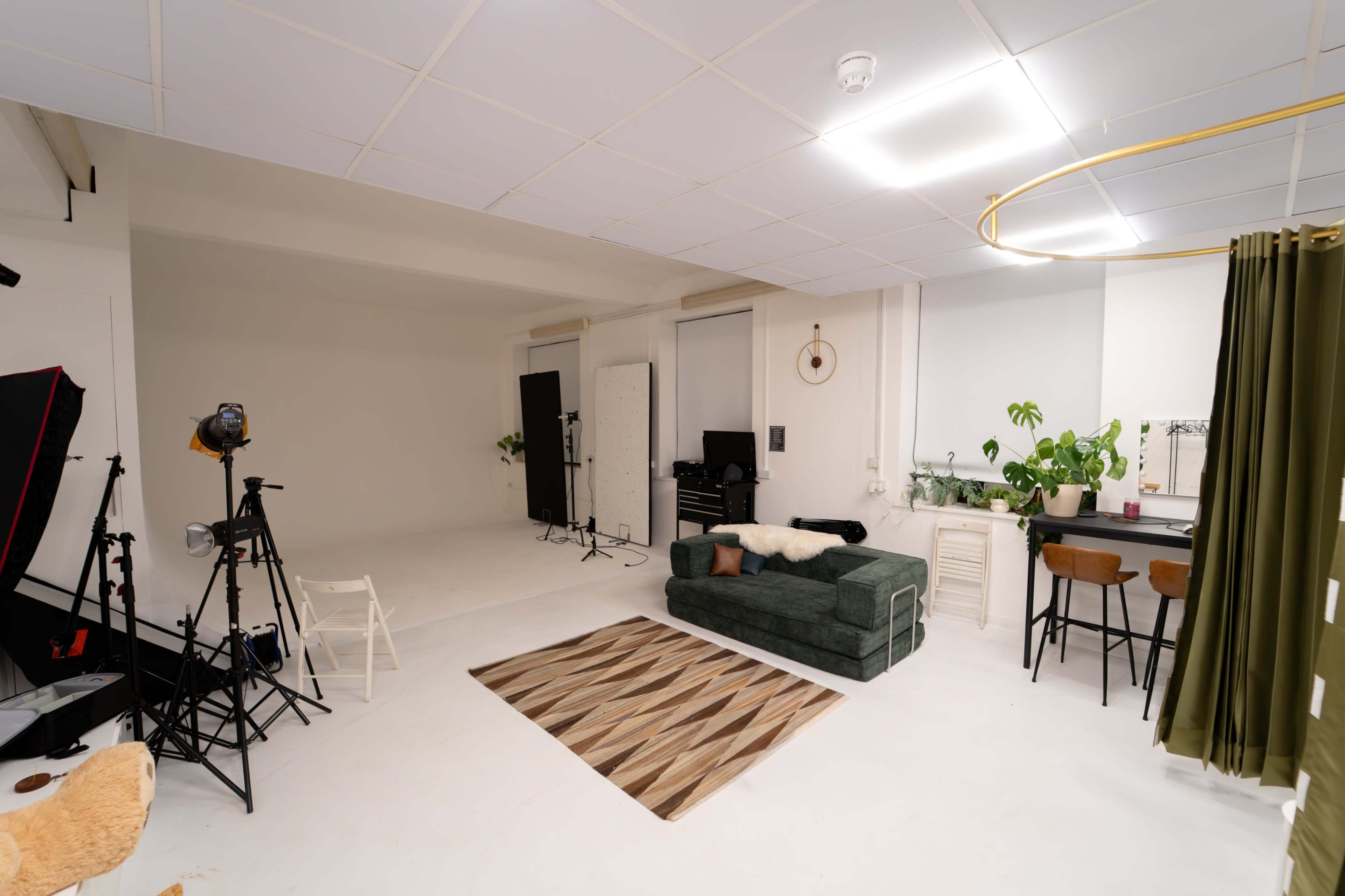 The image shows a spacious photography studio featuring a green sofa, a rug, lighting equipment, and plants in a well-lit environment.