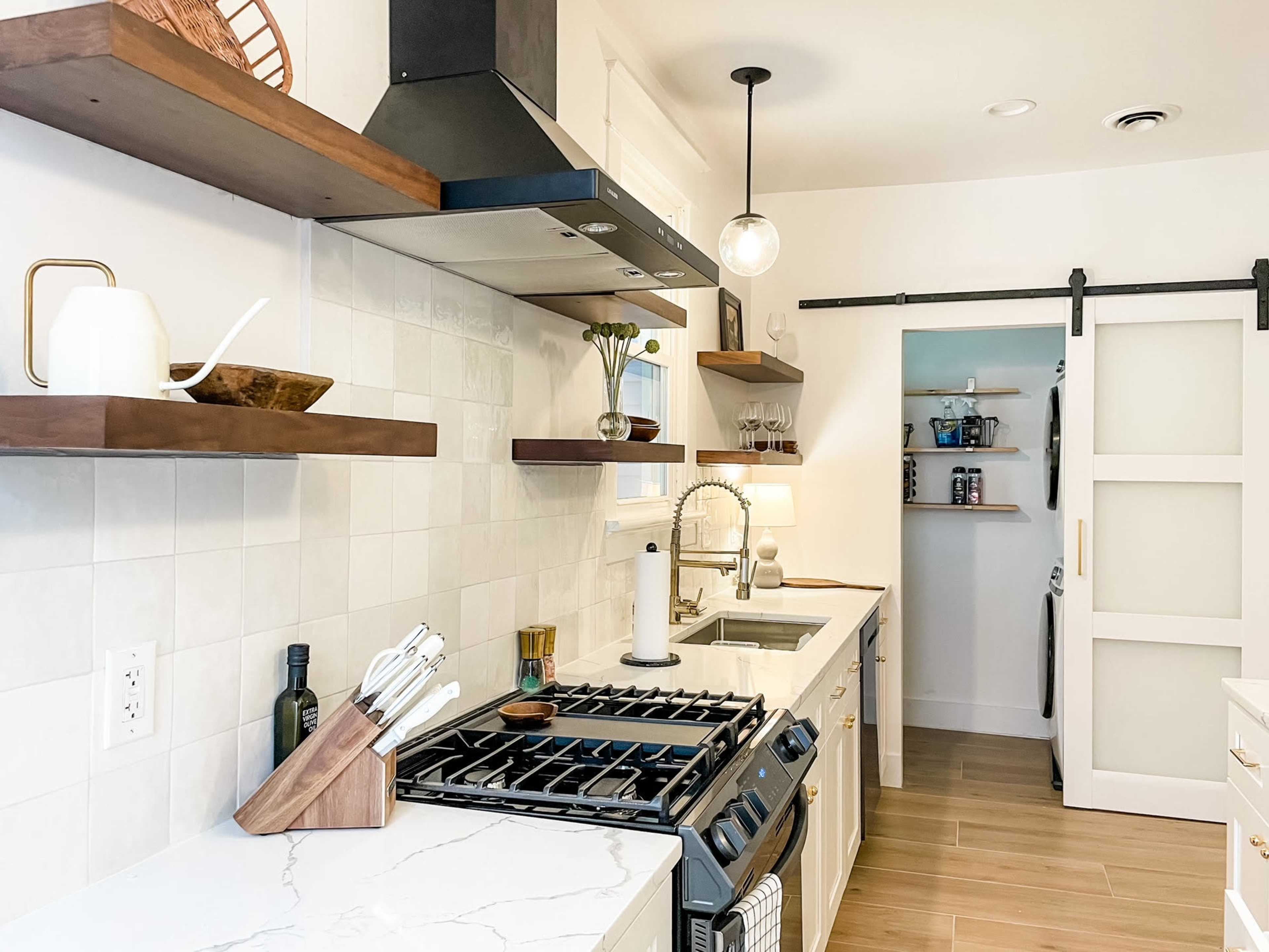 A modern kitchen features white cabinetry, wooden open shelves, a gas stove, and a sliding door to a pantry.