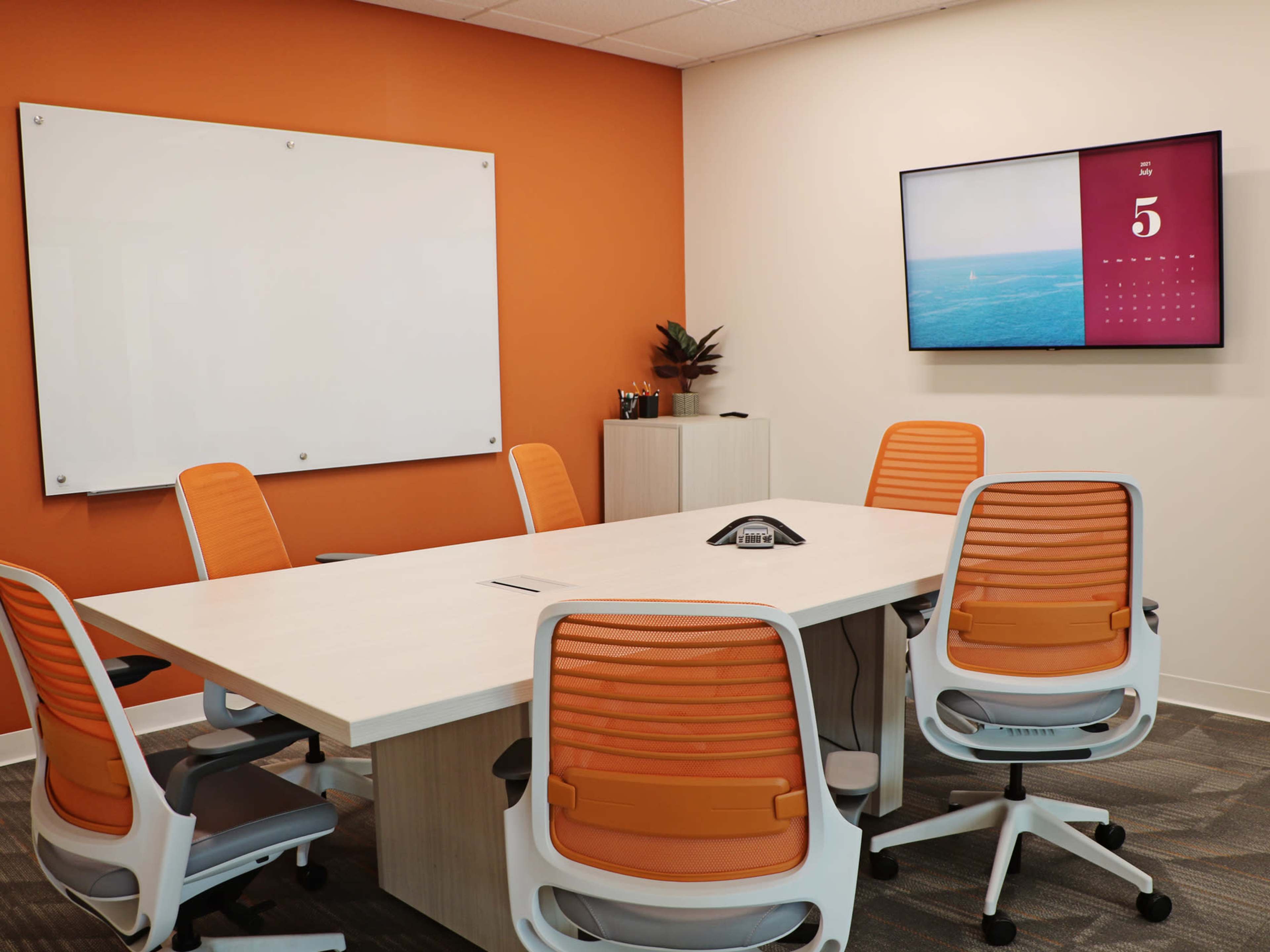 A modern conference room features a whiteboard, a wall-mounted TV displaying a calendar, a central table, and several orange and white chairs.