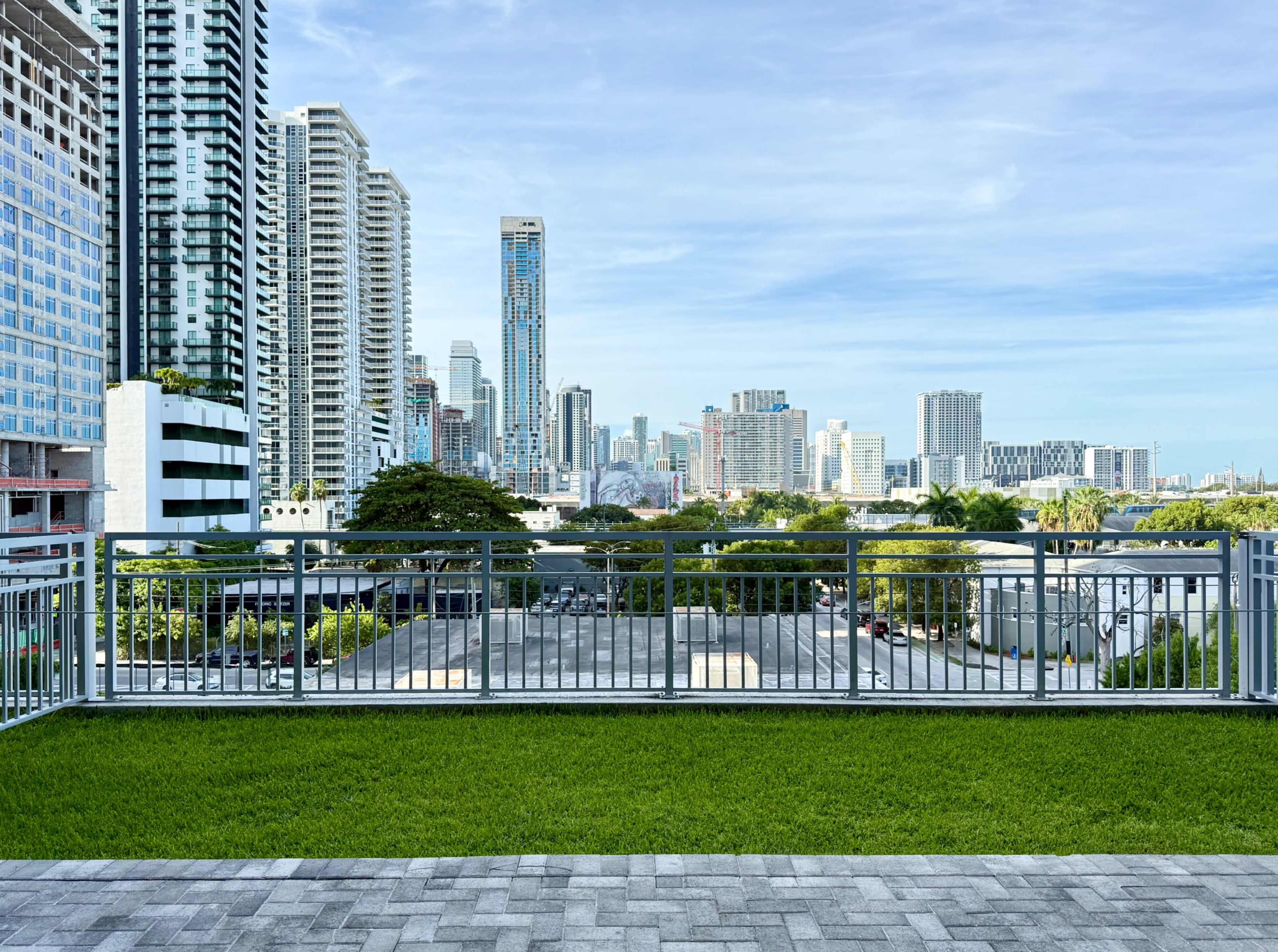 The image shows a view of a city skyline with tall buildings, green grass in the foreground, and a blue sky overhead.