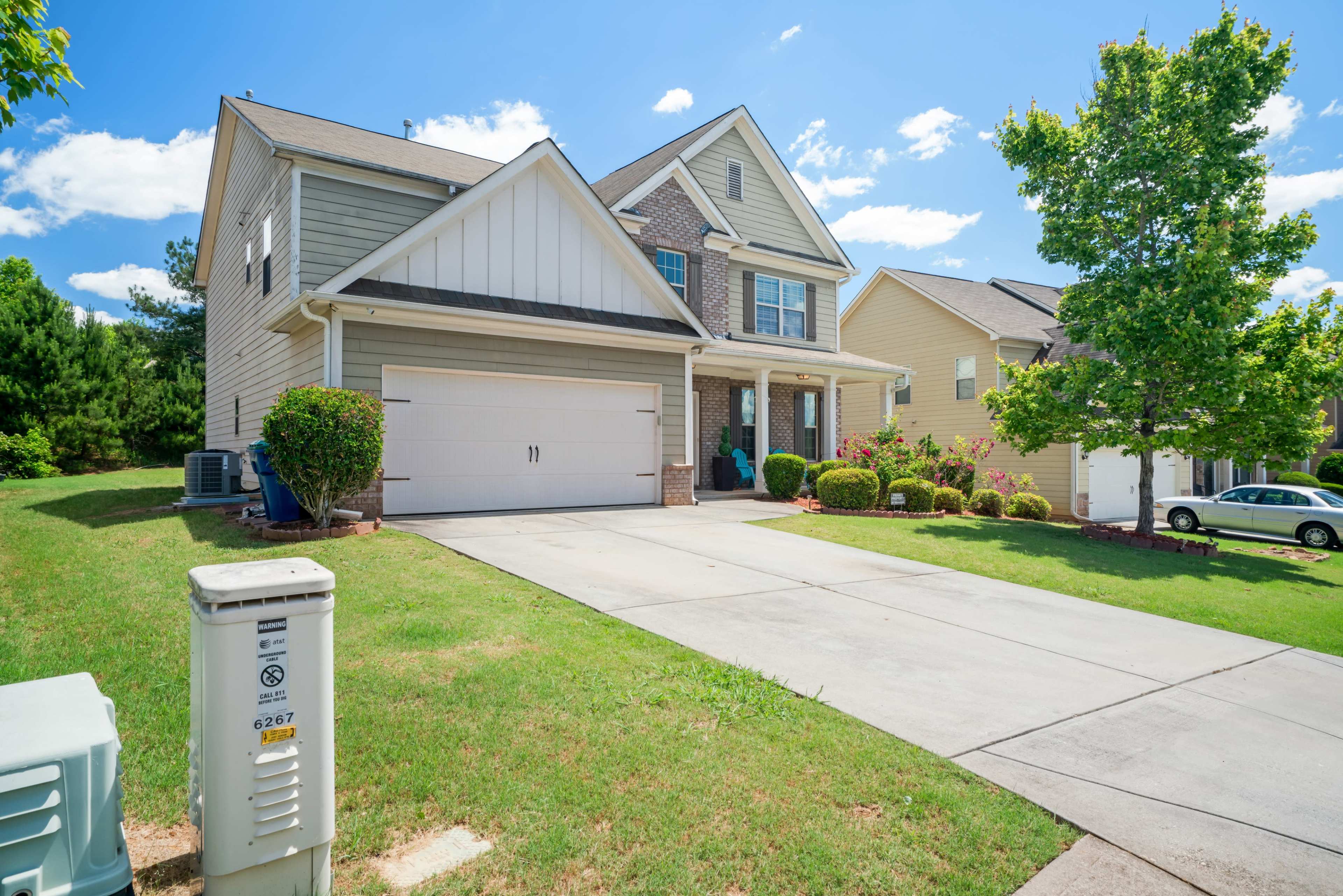 The image shows a two-story house with a driveway, neatly landscaped lawn, and nearby homes under a clear blue sky.