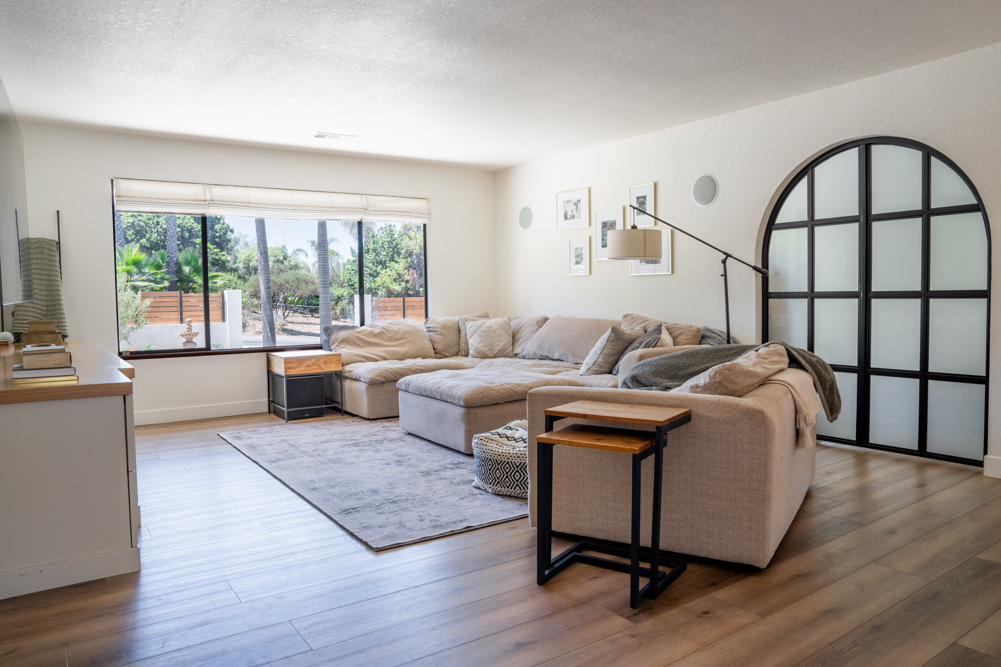 The image shows a bright living room featuring a large sectional sofa, a coffee table, and an arched glass door leading to an outdoor area.