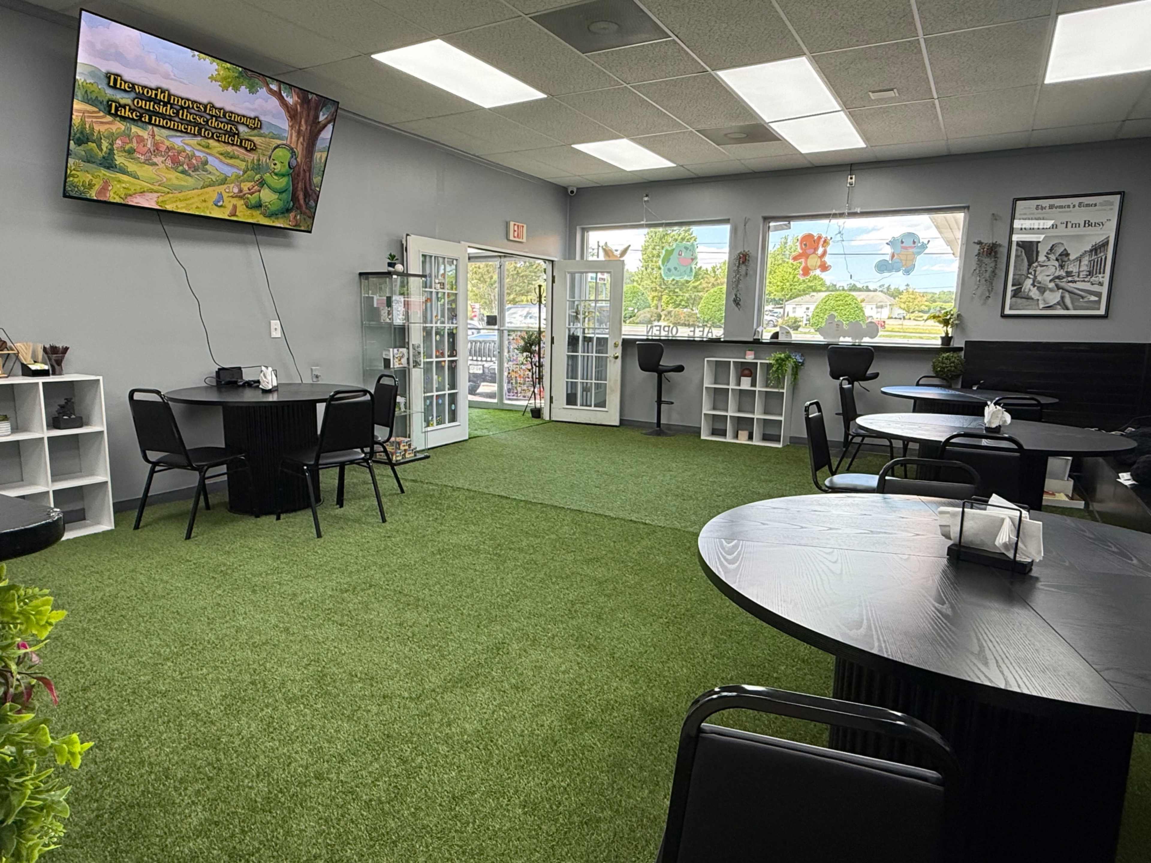 The image shows a cozy indoor café setting with artificial grass flooring, round black tables, and a large TV screen displaying colorful graphics.