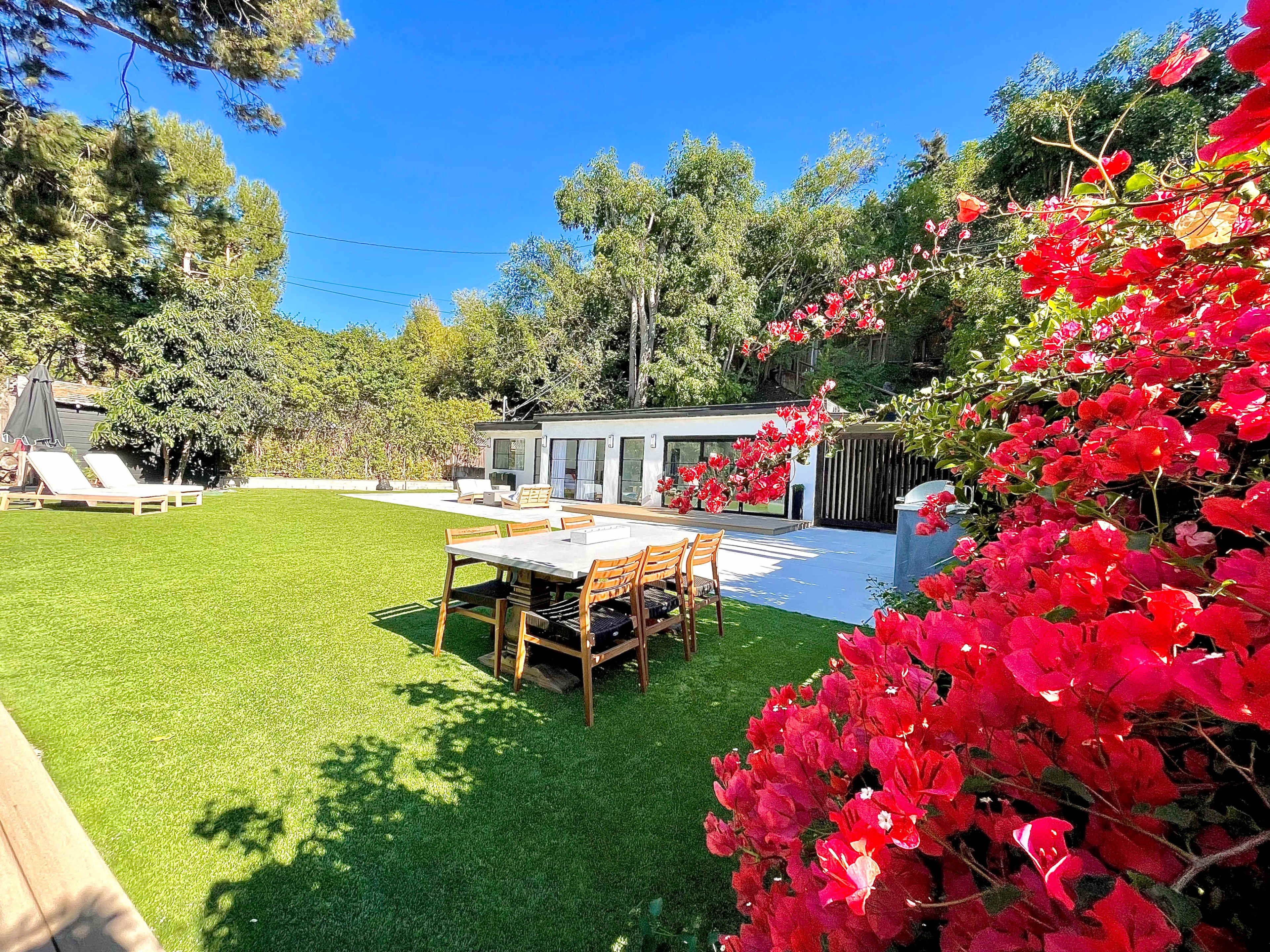A well-maintained outdoor space features a grassy area, a dining table with chairs, and a modern house surrounded by greenery and flowering plants.
