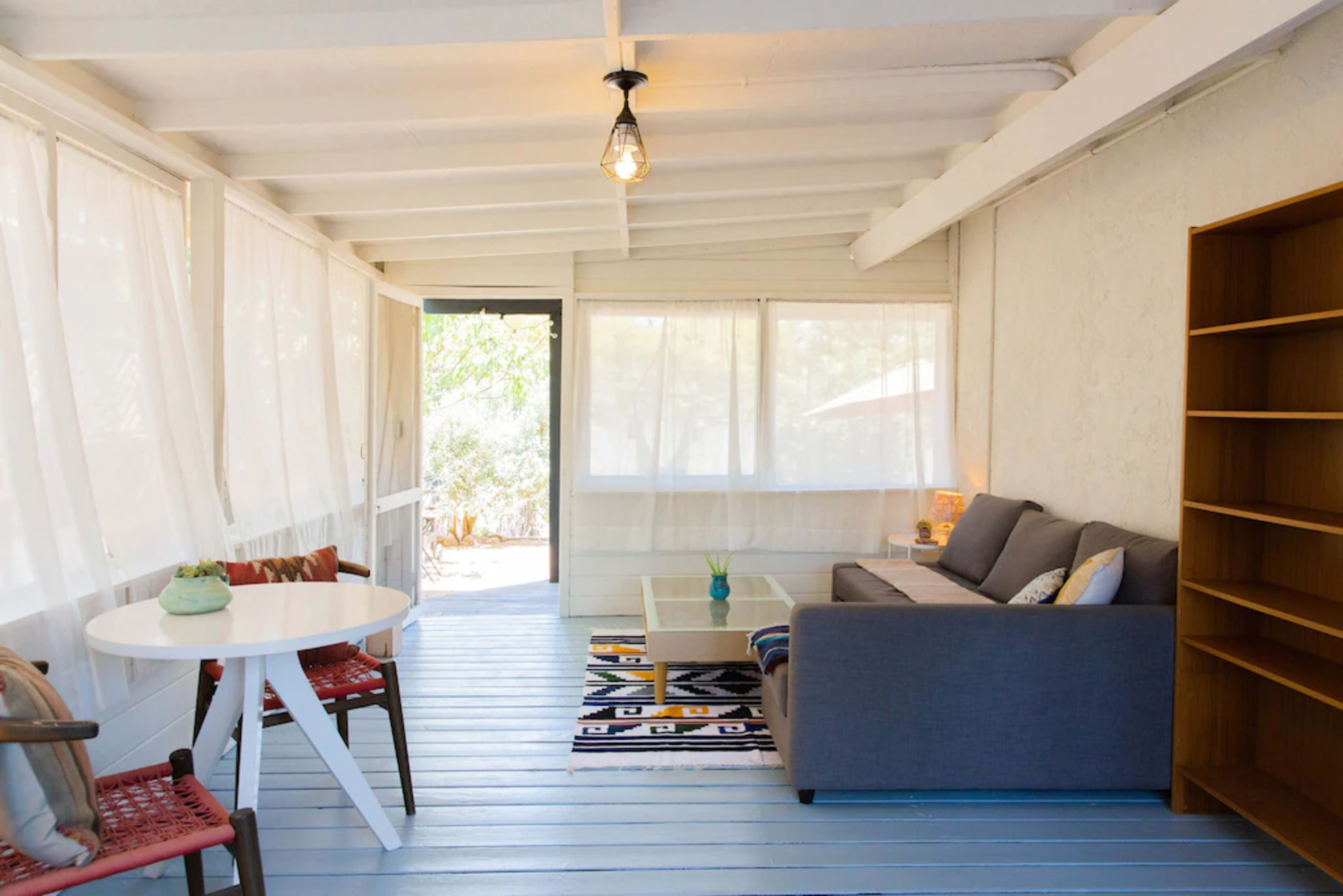 A sunlit sunroom with white walls, a gray sofa, a small round table, and a wooden bookshelf.
