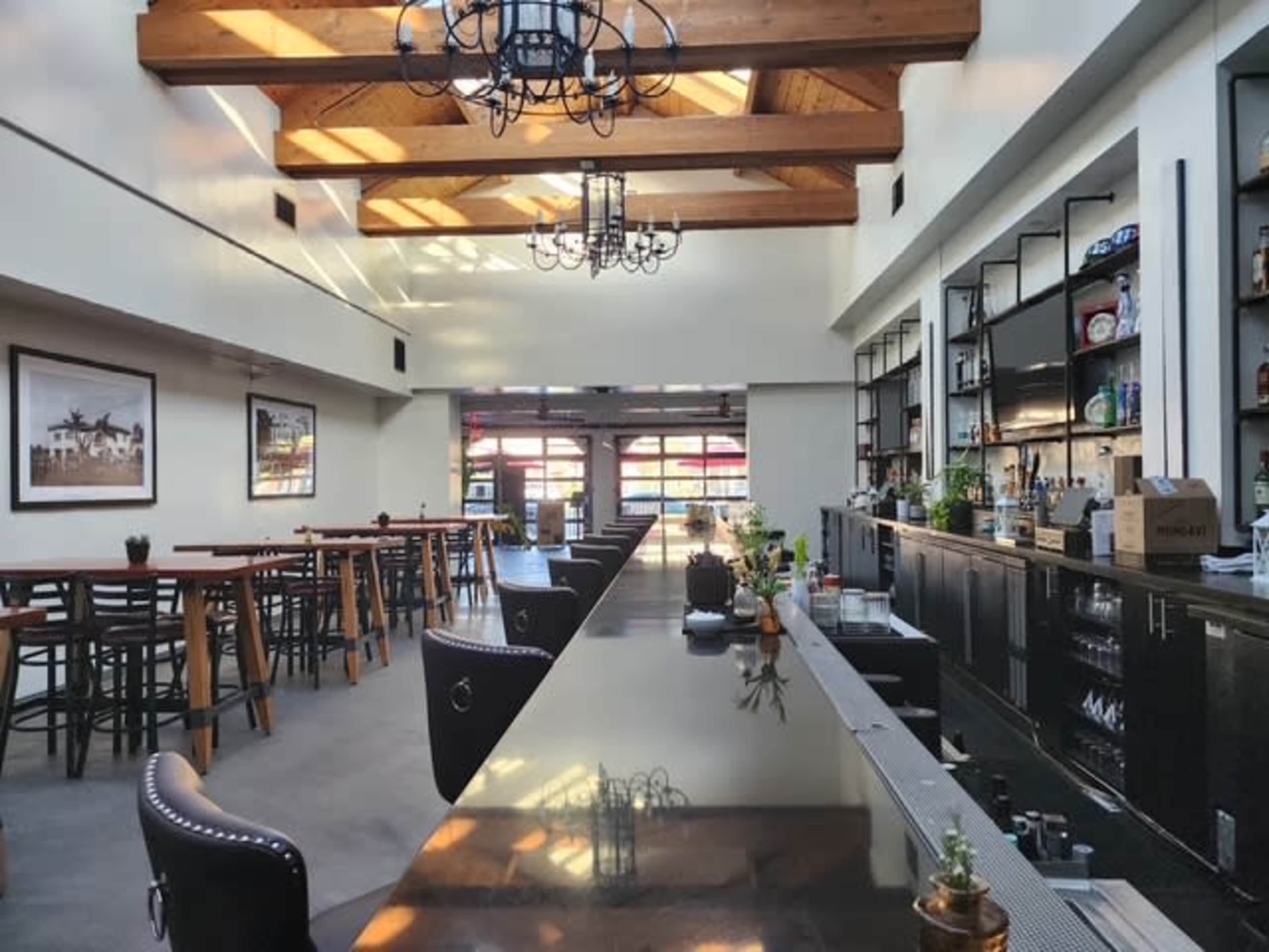 The image shows an interior view of a modern bar and dining area featuring wooden beams, a long bar counter, and several tables lined along the walls.