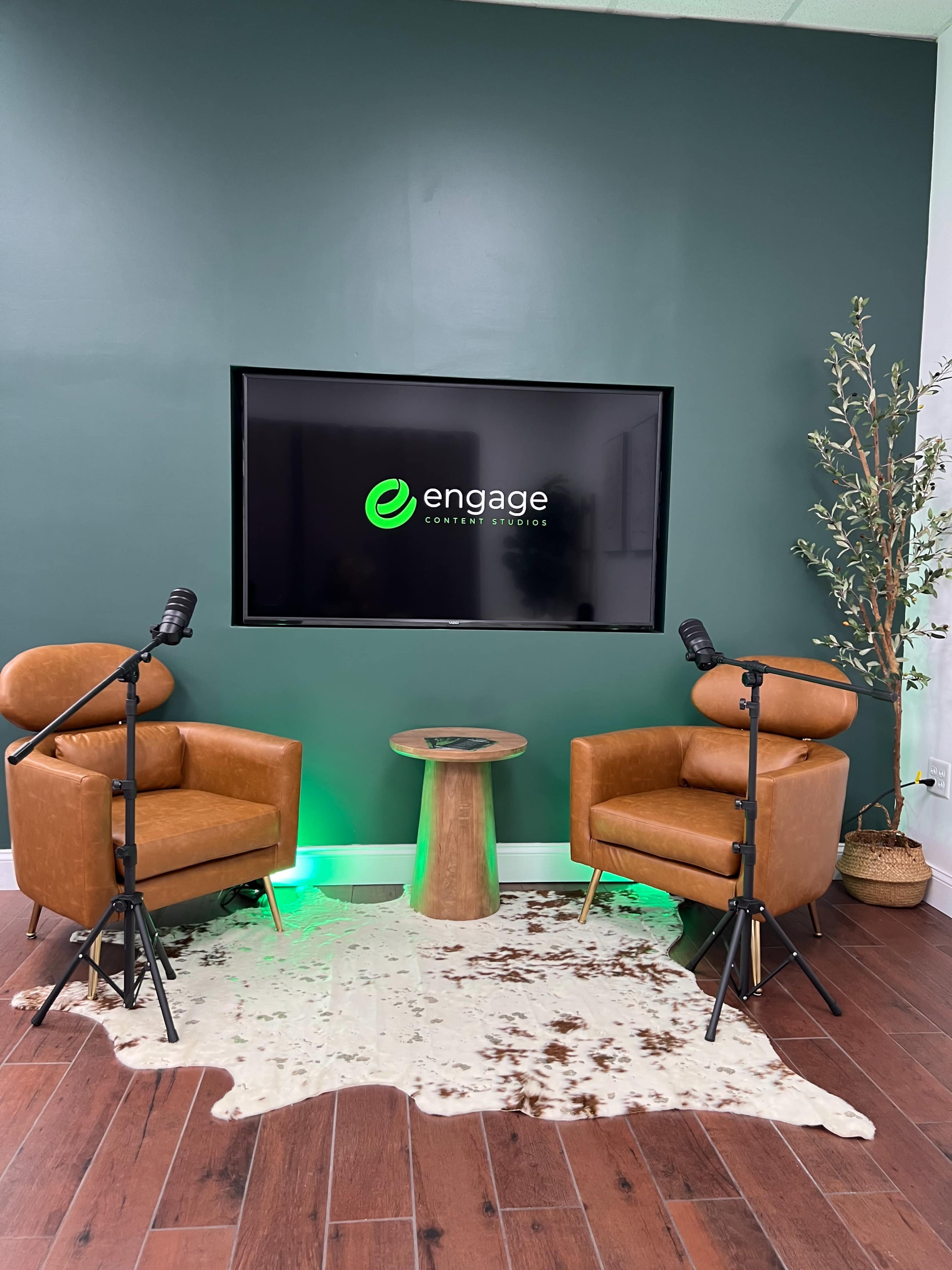 A studio setup featuring two brown leather chairs, a wooden side table, and a TV mounted on a green wall with the logo "engage" displayed.