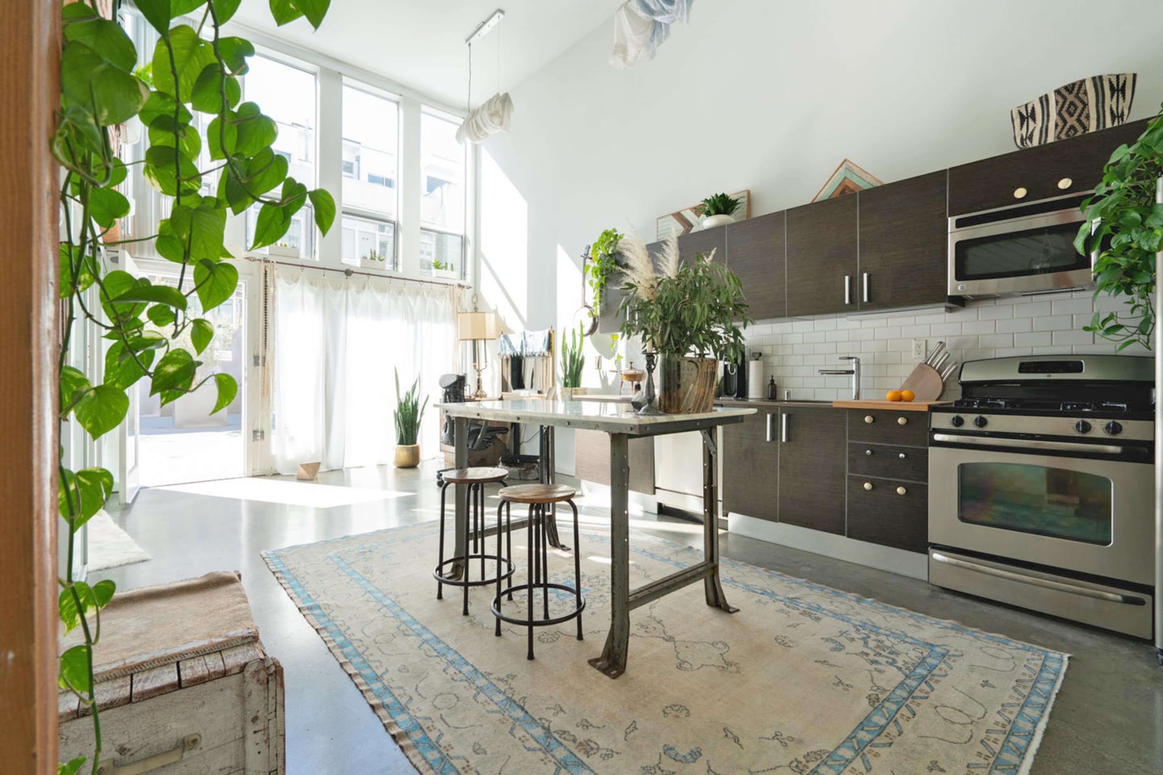The kitchen features a central island with two stools, surrounded by modern cabinetry, plants, and large windows that allow ample natural light.