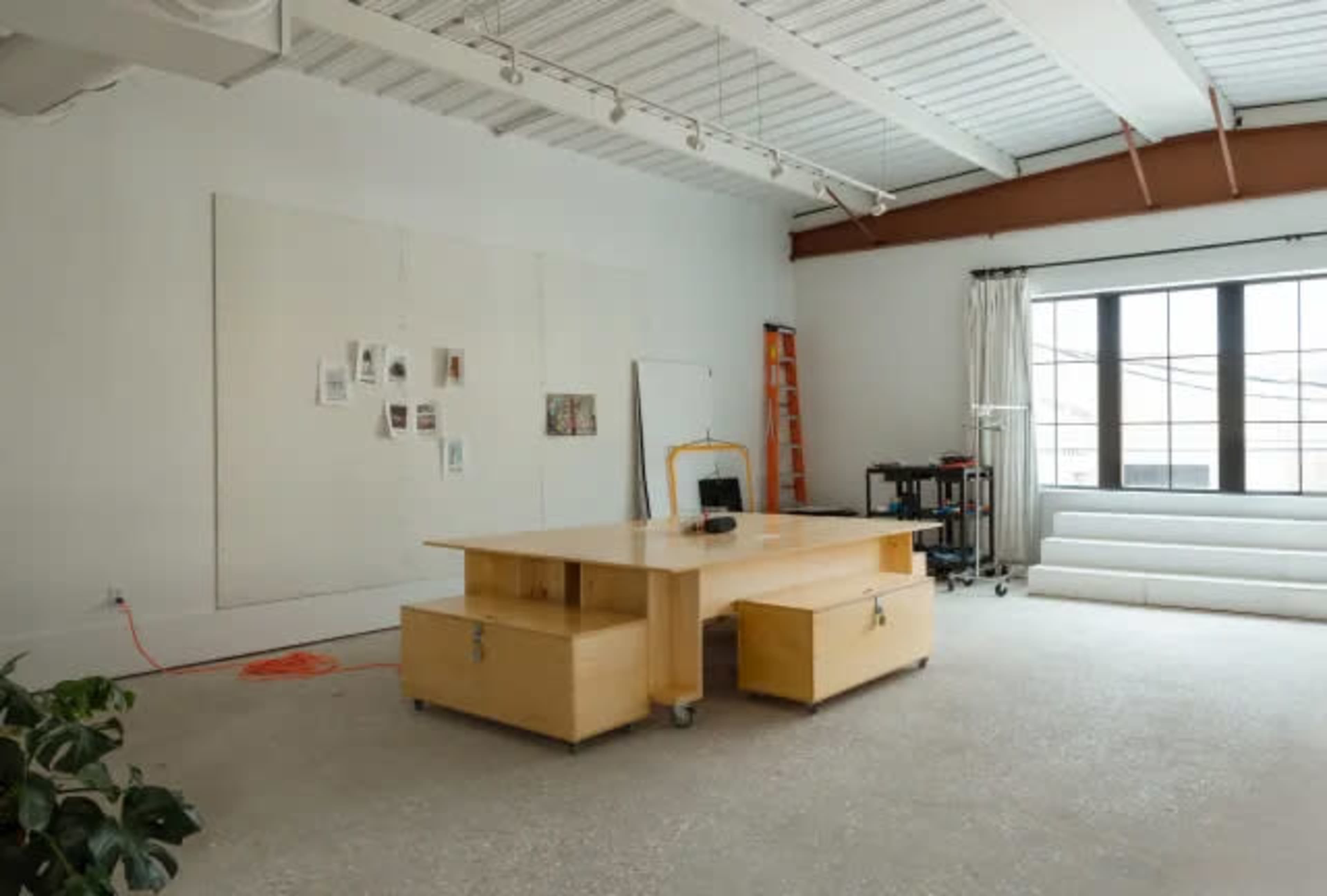The image shows a spacious, minimalist office or studio featuring a central wooden desk, a few pinned documents on the wall, a ladder, and white furniture arranged by a large window.