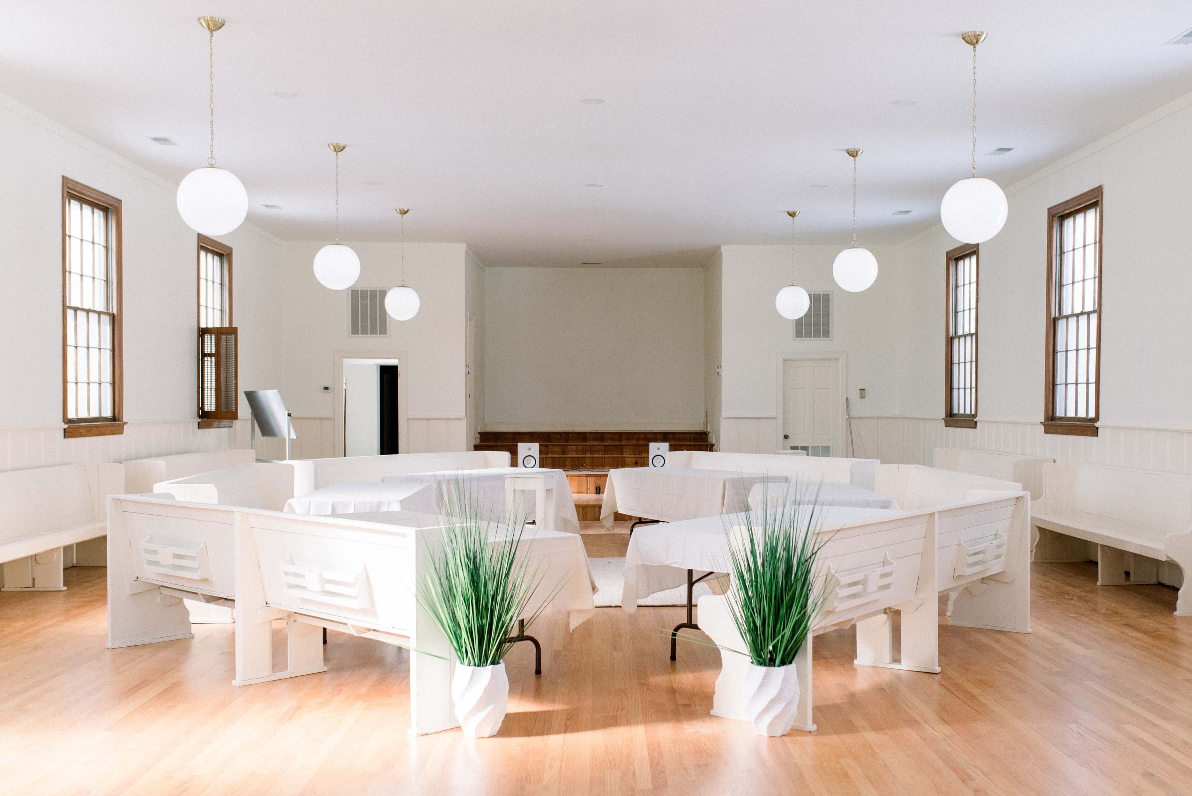 A spacious, well-lit room with white walls, featuring round tables arranged in a circular layout and potted plants in white coverings.