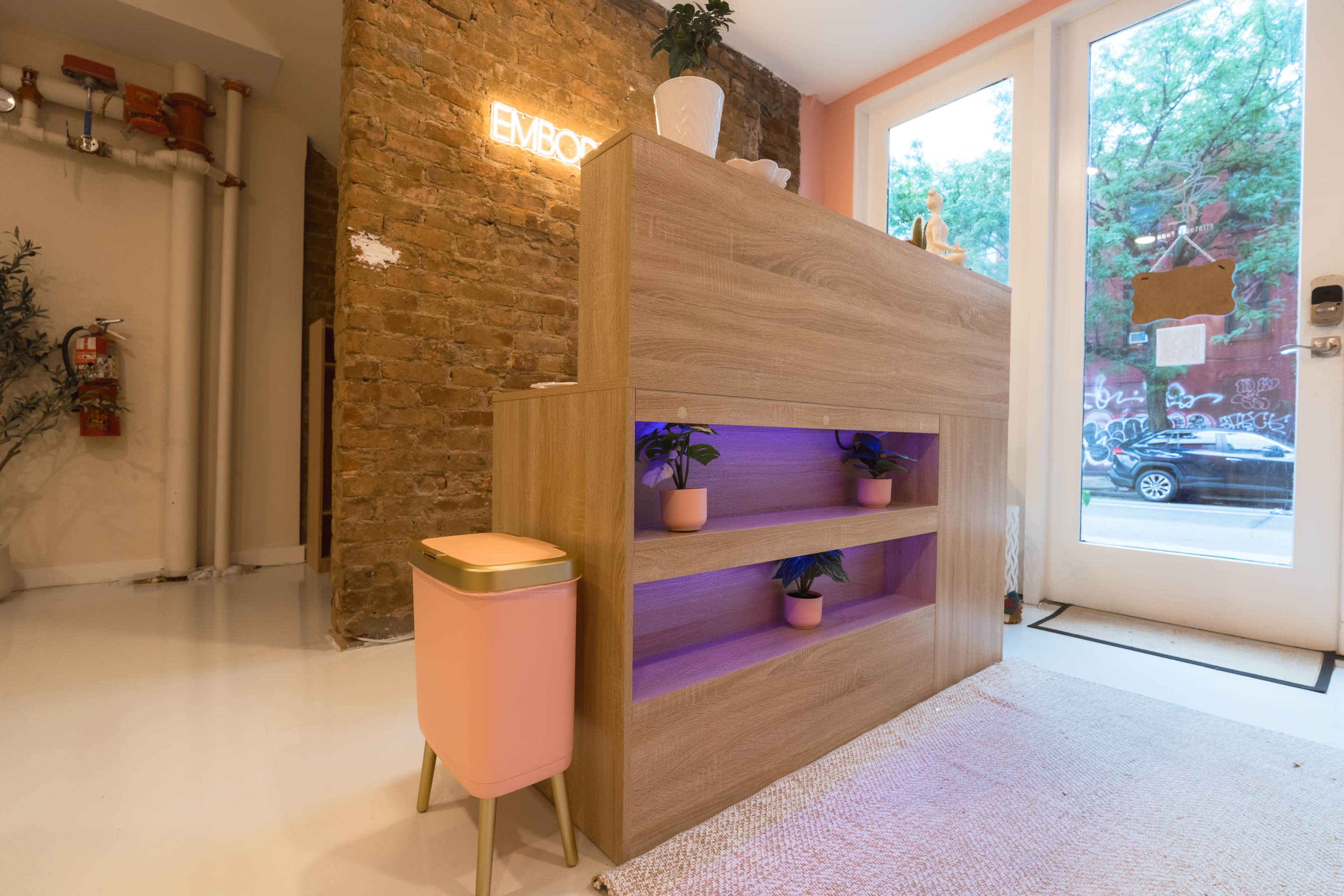 The image shows a modern reception desk with wooden panels and decorative plants, situated in a well-lit entryway with a brick wall and large windows.