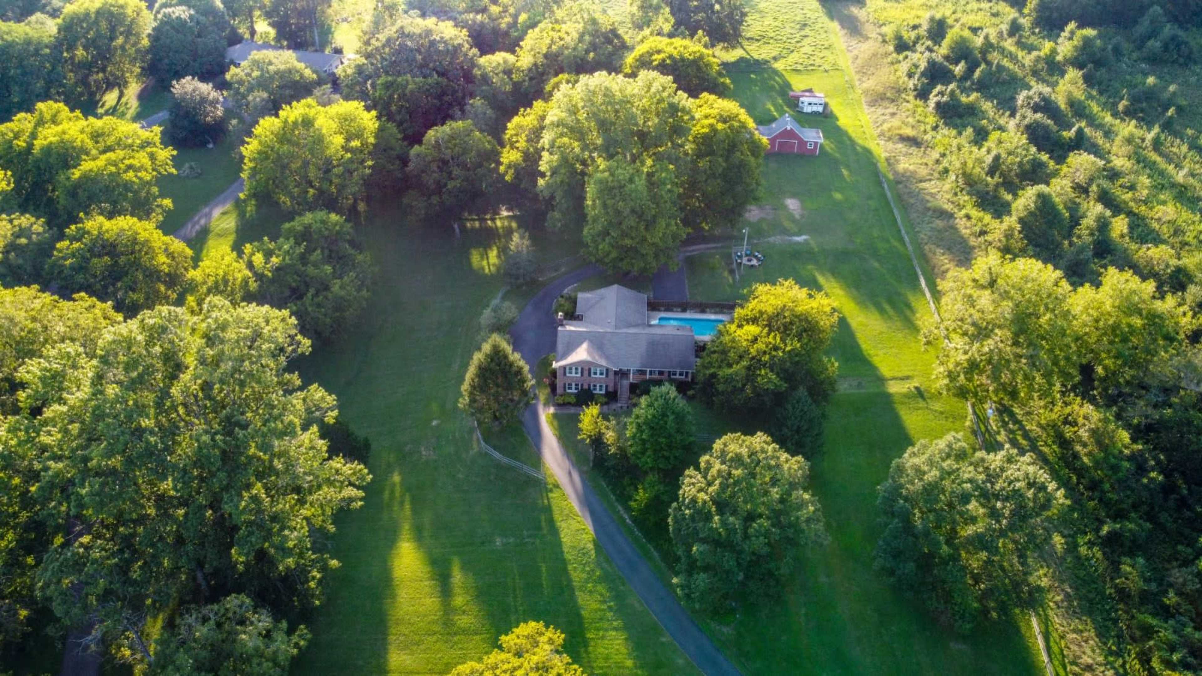 A large house surrounded by trees and grass, with a swimming pool and barn visible in the yard.