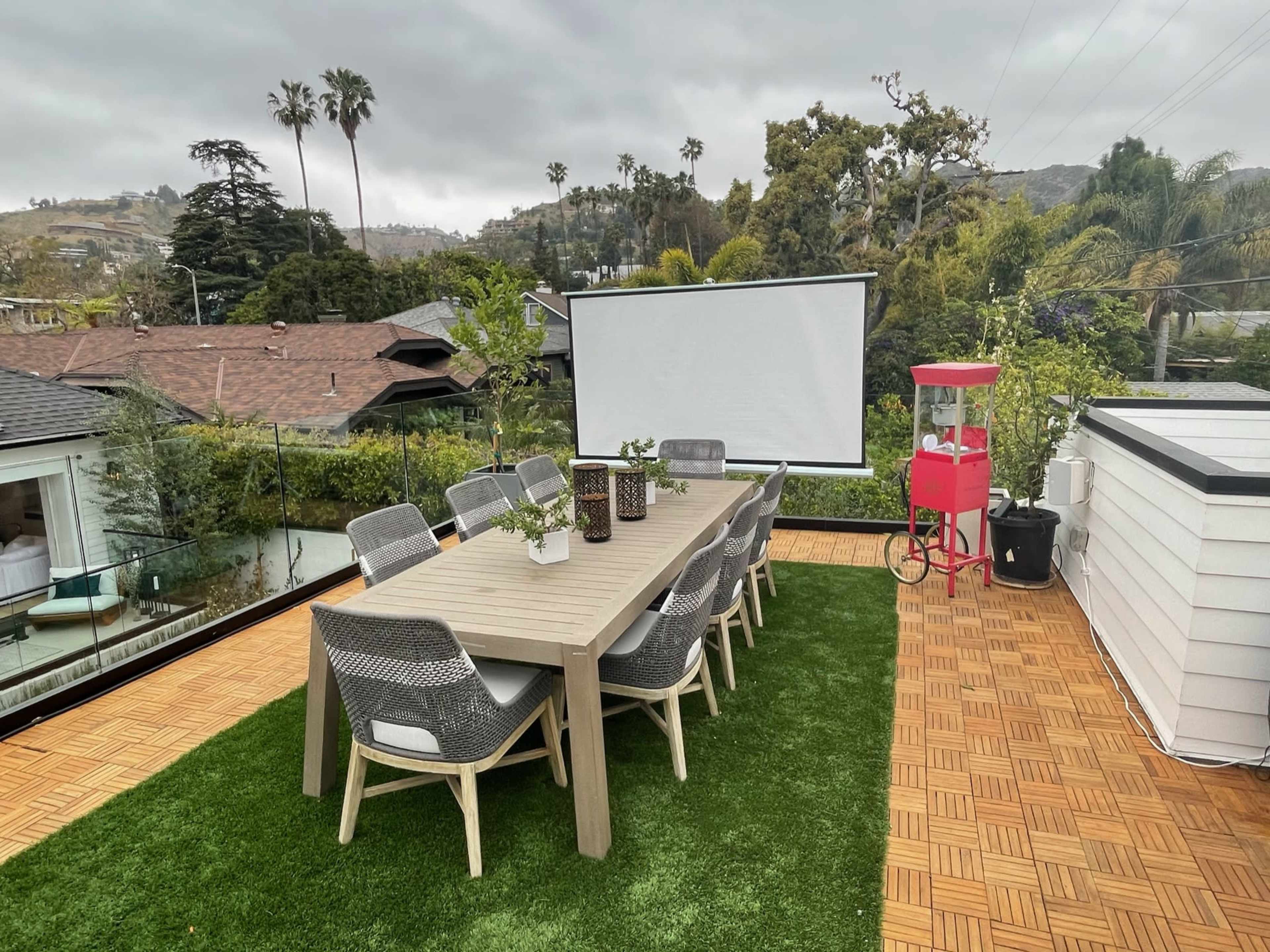 A rooftop terrace features a long wooden dining table surrounded by gray chairs, a large screen for presentations, and a red cart in the corner, all set against a backdrop of palm trees and hills.