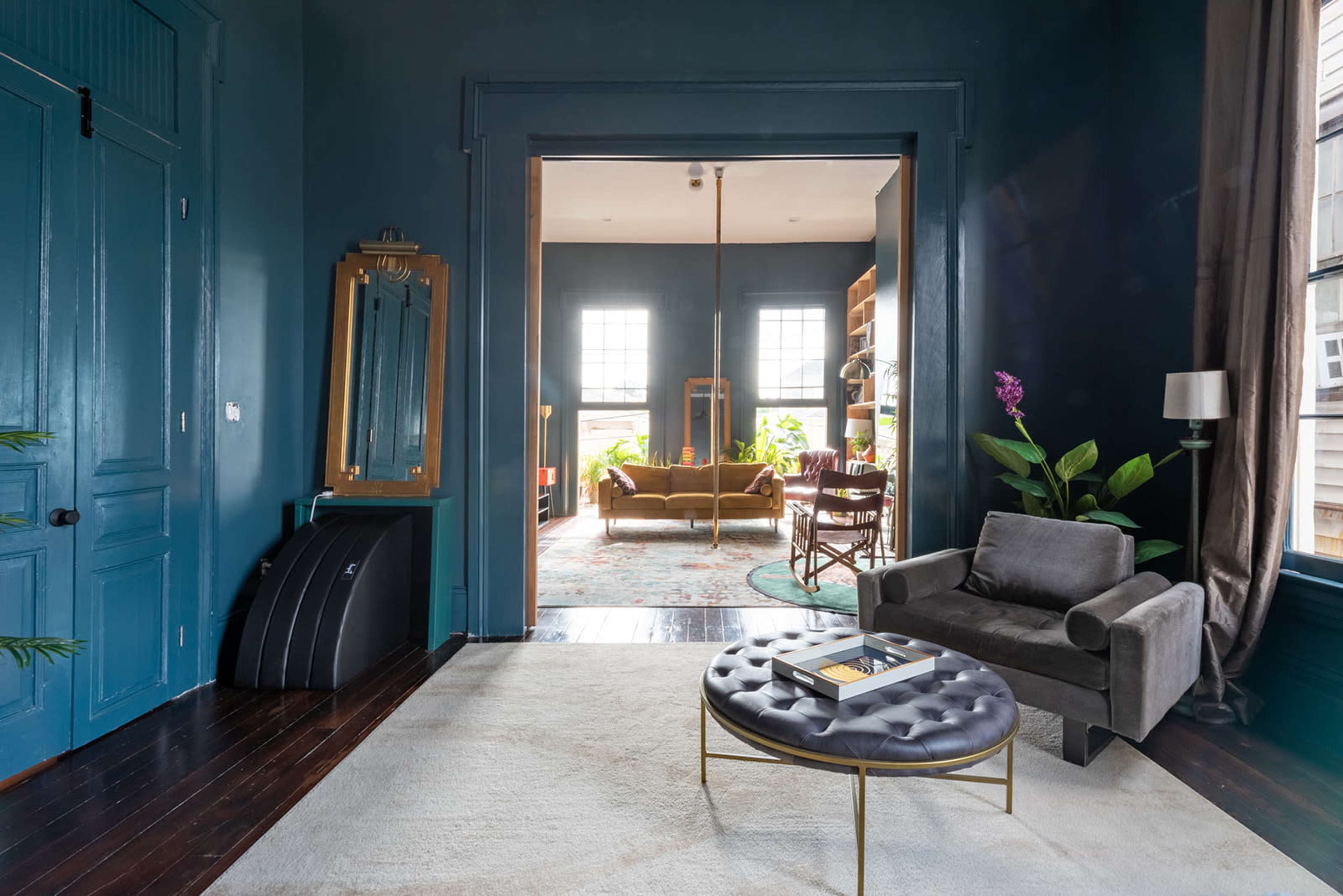 A spacious, modern living room features dark teal walls, large windows, and a light-colored rug with a round coffee table in the foreground.