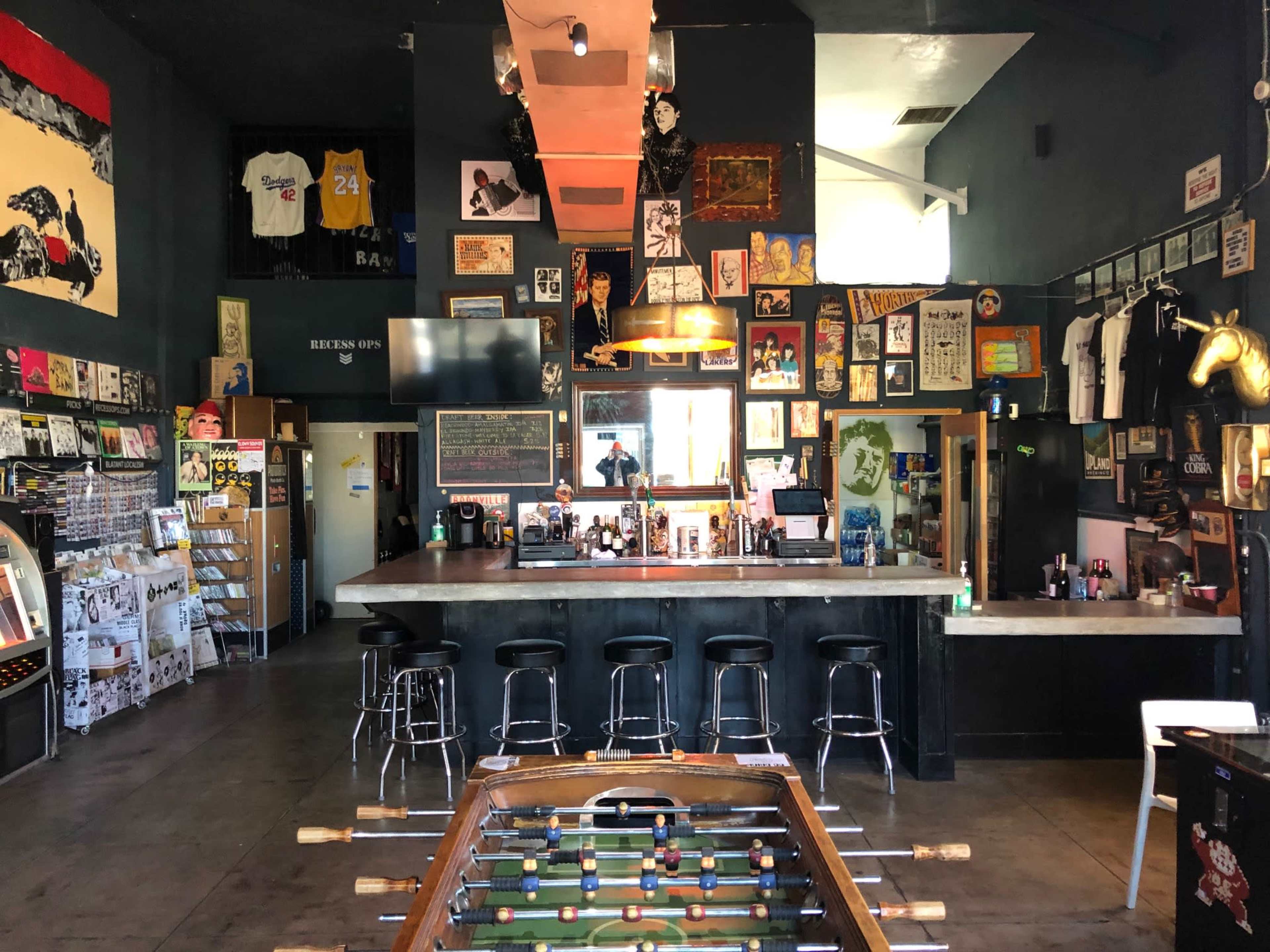 A dimly lit bar with a foosball table in the foreground, surrounded by various posters and artwork on the walls.