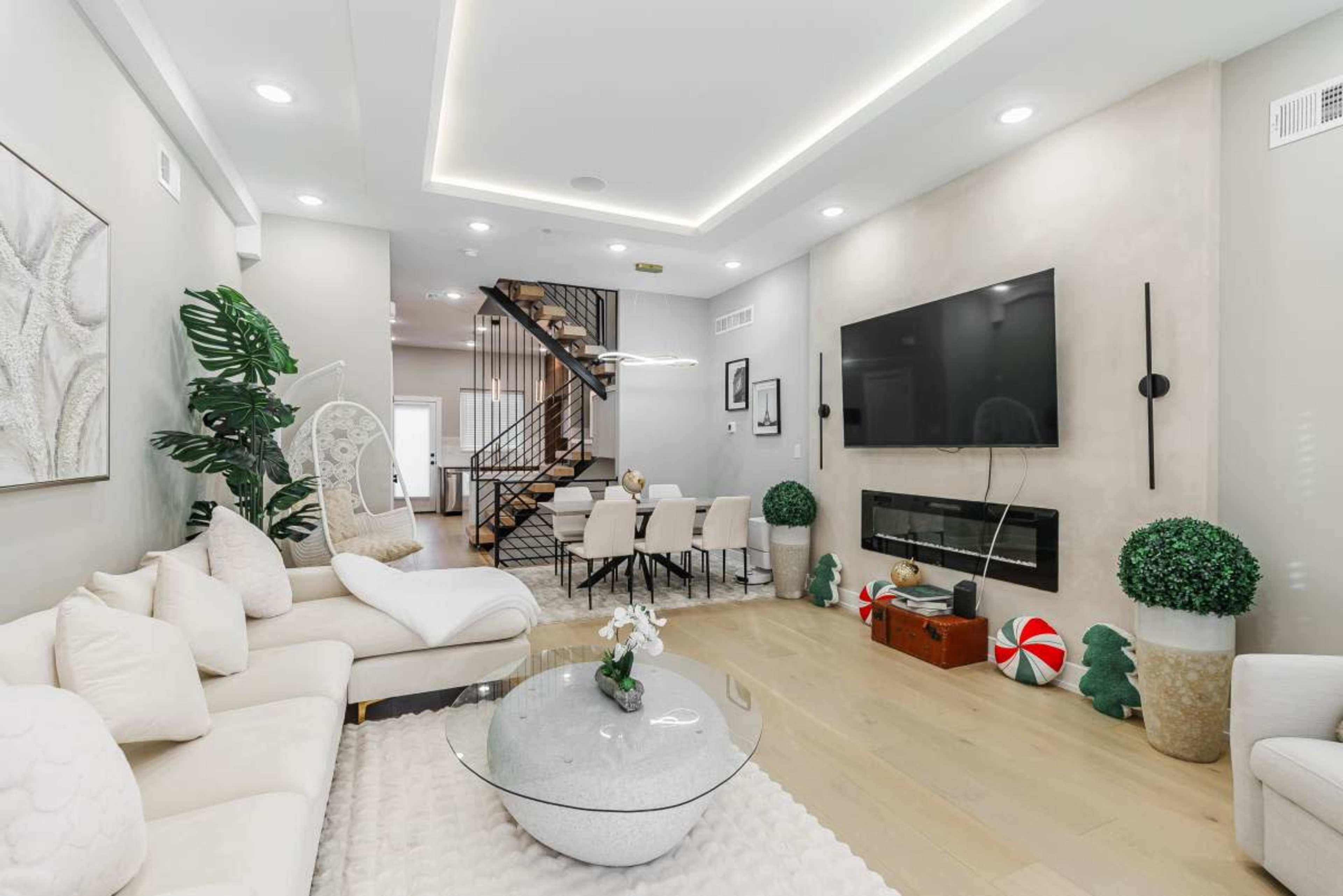 The image shows a modern living room with a white sectional sofa, a glass coffee table, and a large television mounted on a light gray wall, adjacent to a dining area and a staircase.