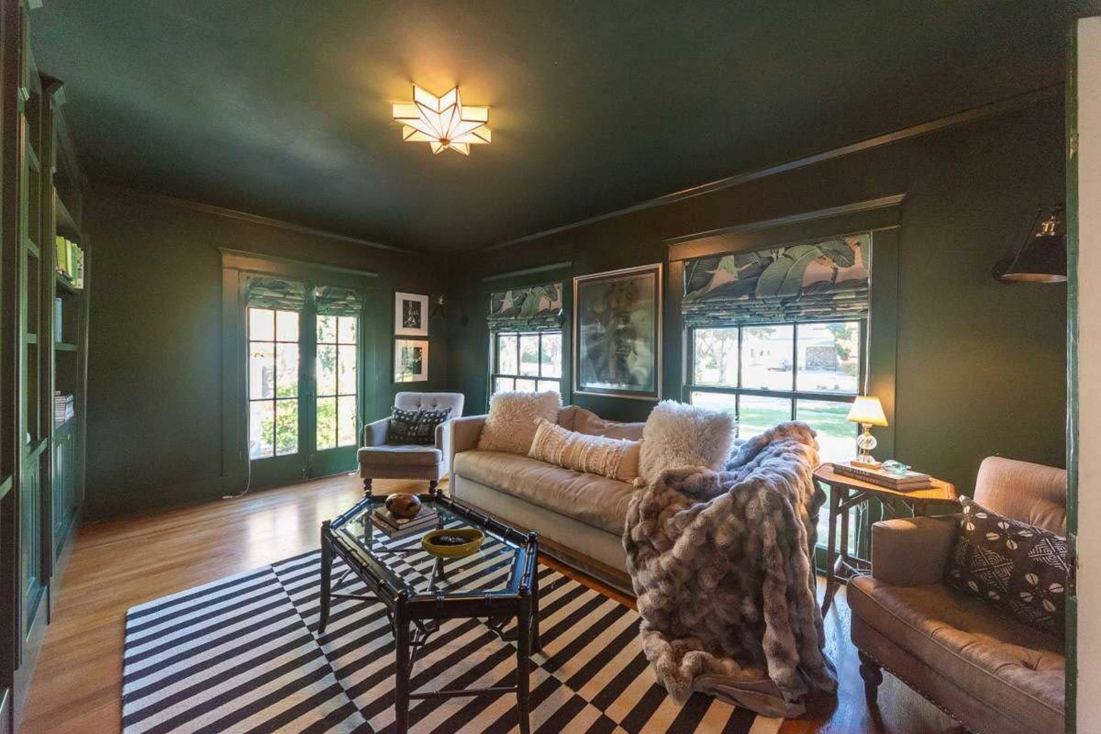 The room features dark green walls, large windows with patterned shades, a beige sofa with decorative pillows, and a geometric black-and-white rug spread across the wooden floor.