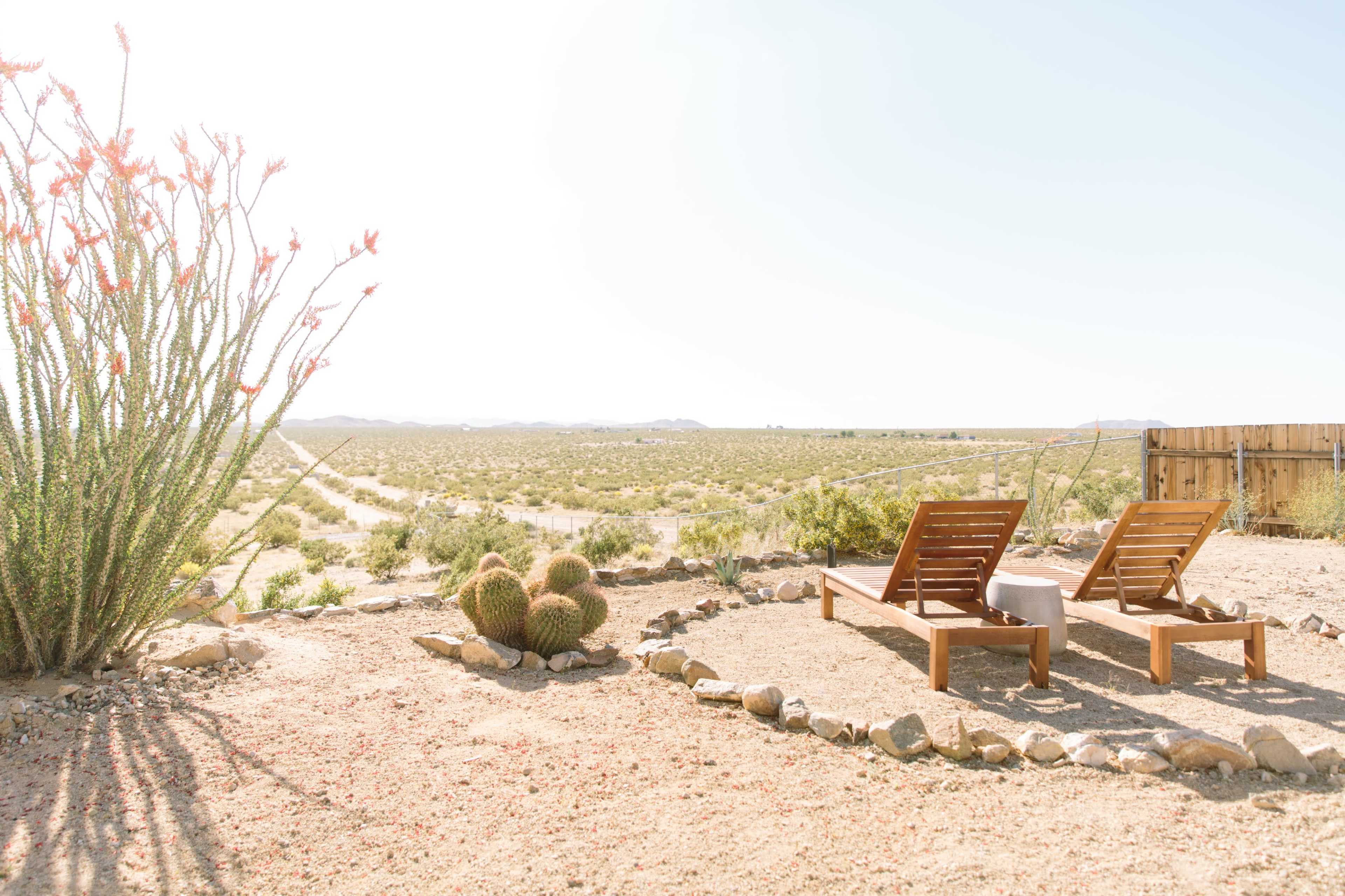 Two wooden lounge chairs are positioned on a gravel area surrounded by rocks, overlooking a vast desert landscape with sparse vegetation and distant mountains.
