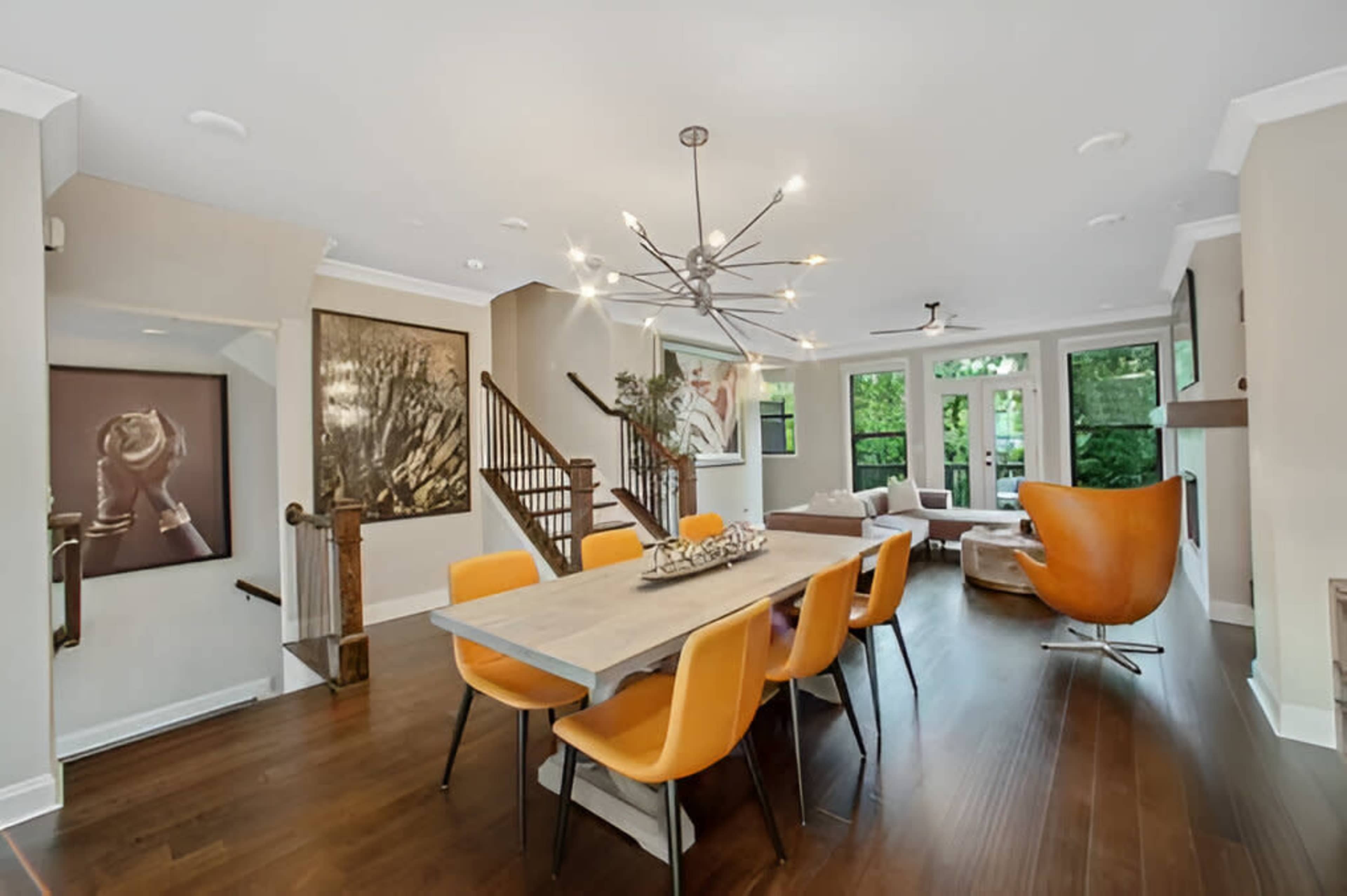 A contemporary dining area featuring a wooden table surrounded by orange chairs, with a staircase and large windows in the background.