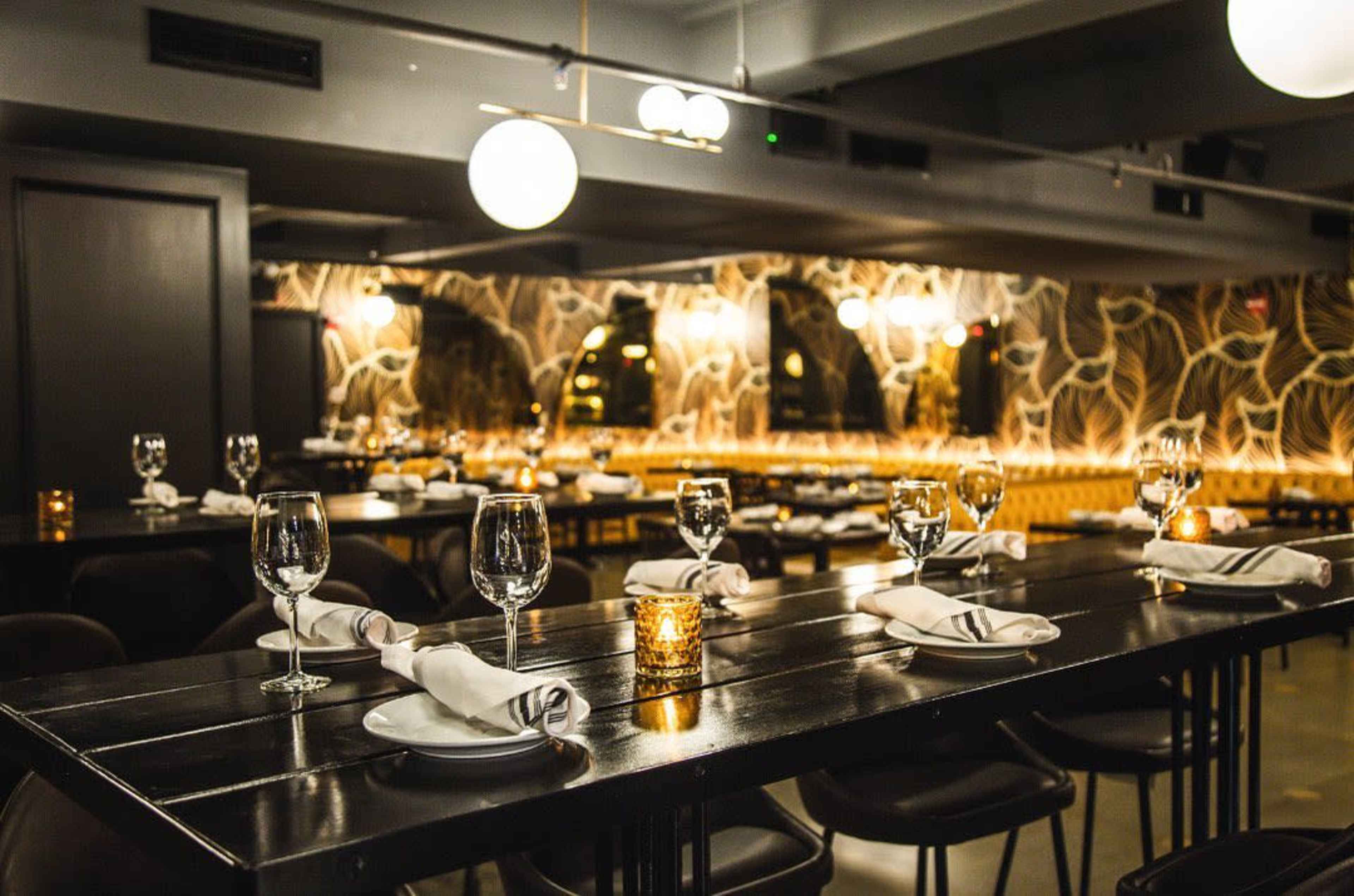 The image shows a modern restaurant interior with long tables set for dinner, featuring glassware and neatly folded napkins, illuminated by ambient lighting.