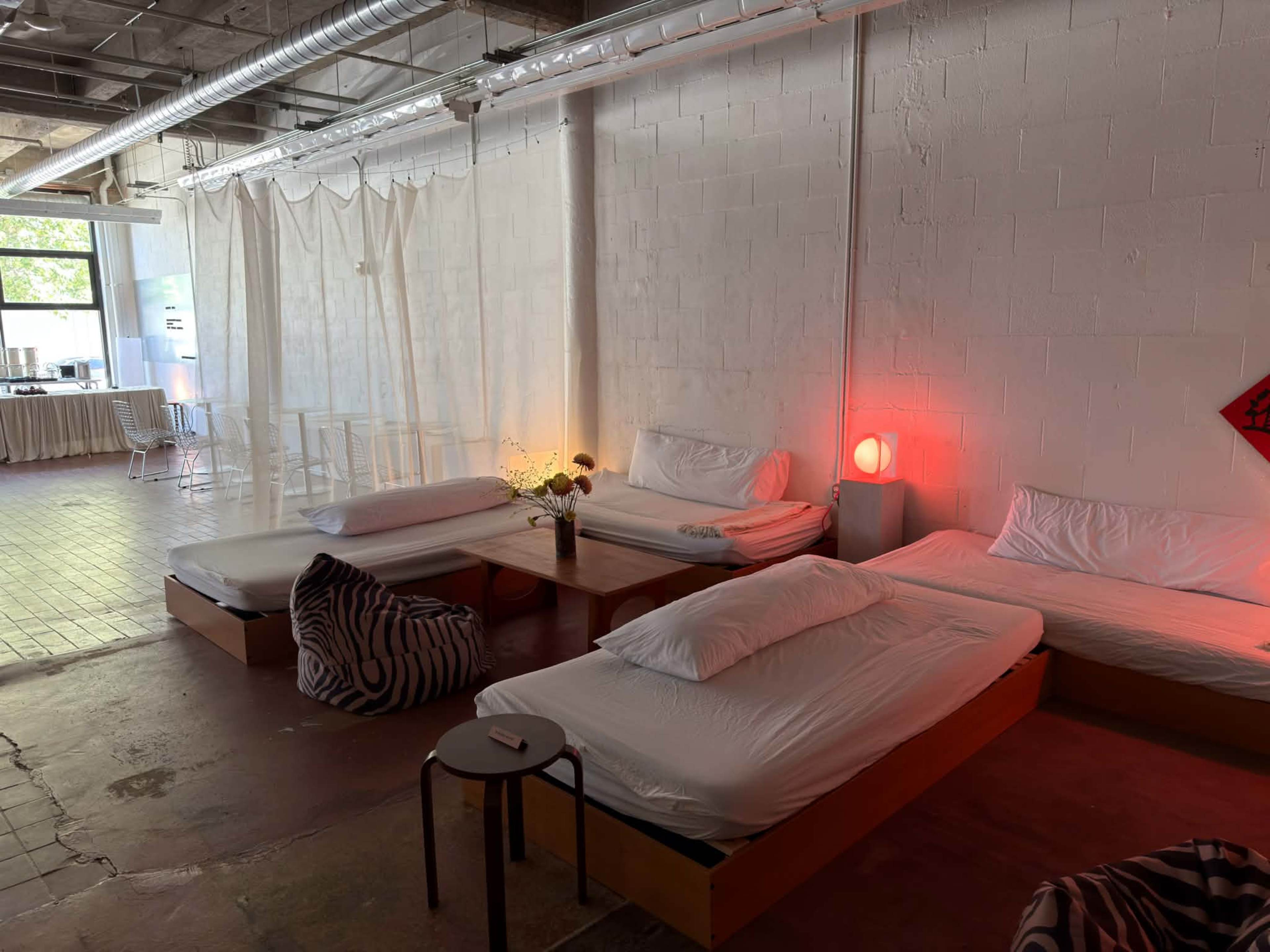 The image shows a spacious room with several unmade beds, a low table, and a small round side table, illuminated by soft red light.