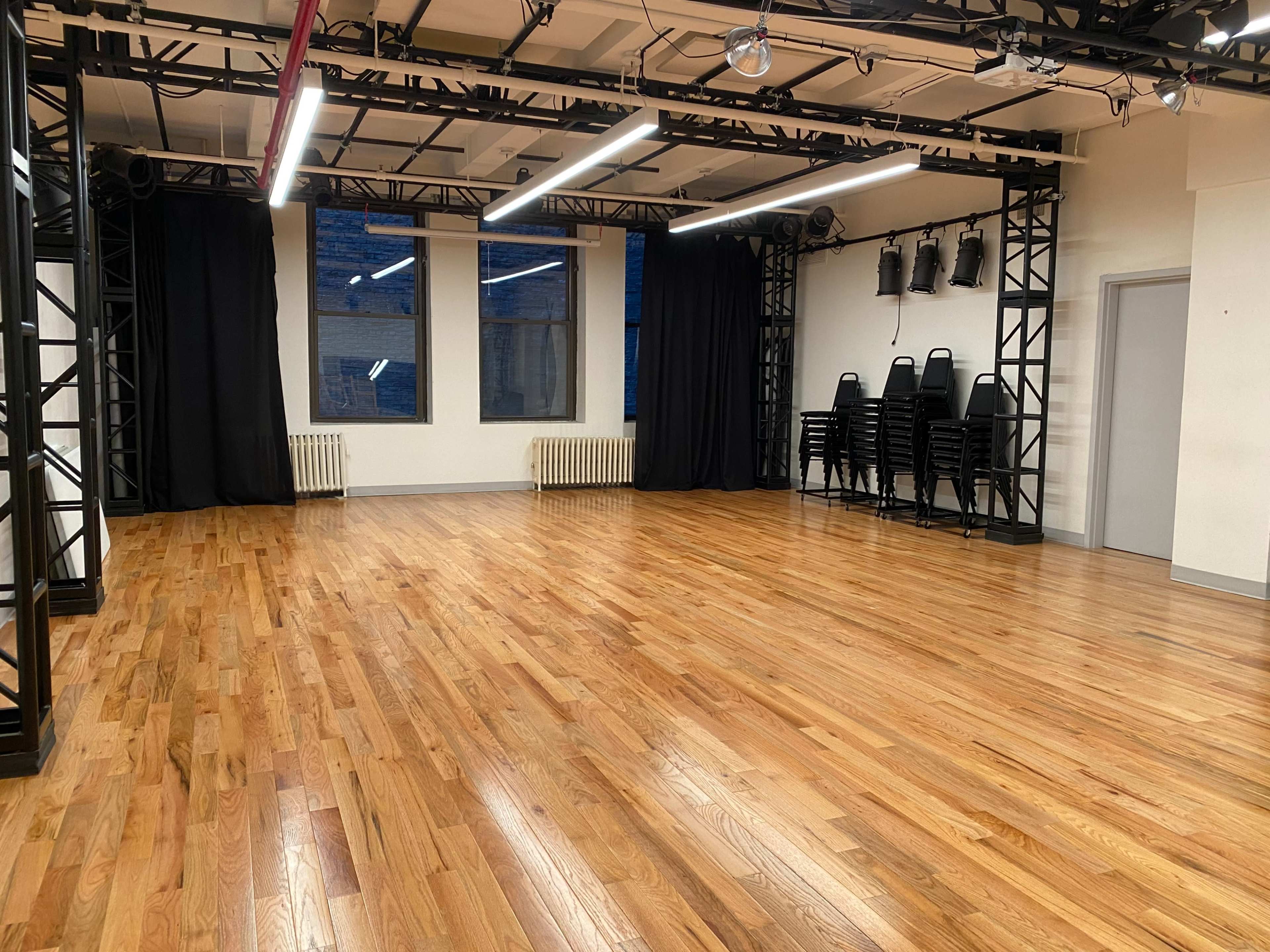 A spacious, empty dance studio with polished wooden floors, large windows, and black curtains, along with stacked chairs in the corner.