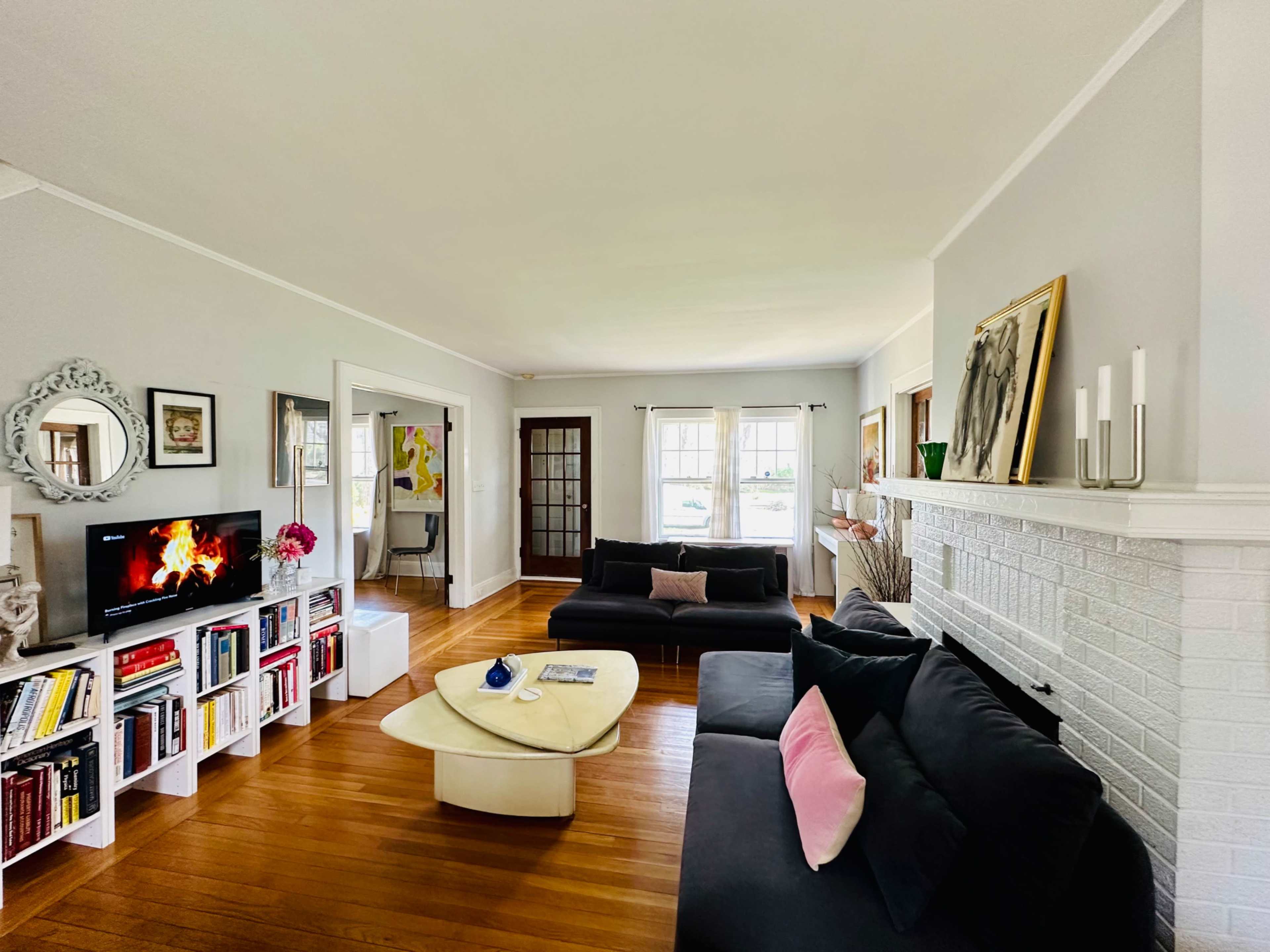 The living room features a pair of black sofas, a circular coffee table, a bookshelf filled with books, and a television displaying a fire, set against light gray walls and hardwood floors.