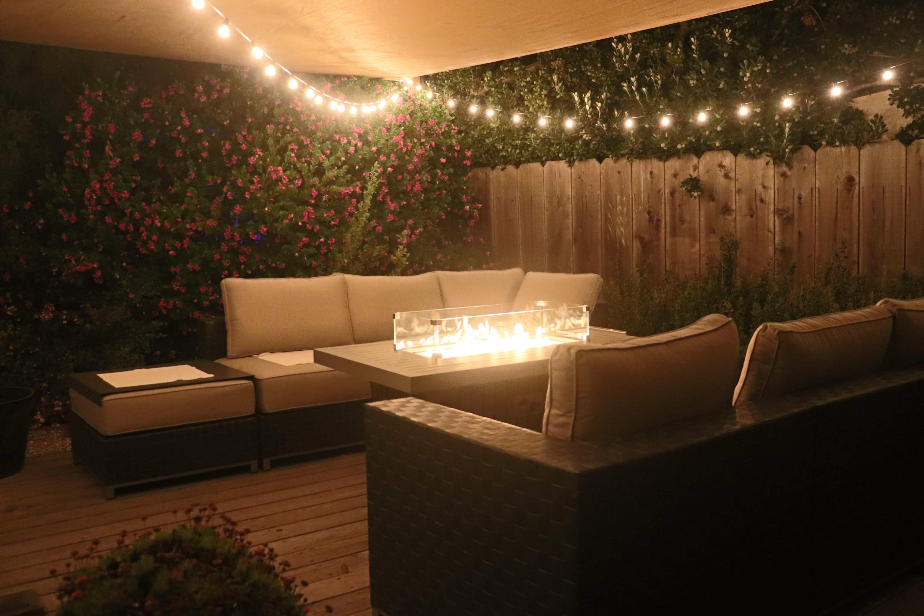 A cozy outdoor seating area features a lit fire table surrounded by cushioned couches under string lights, with foliage in the background.